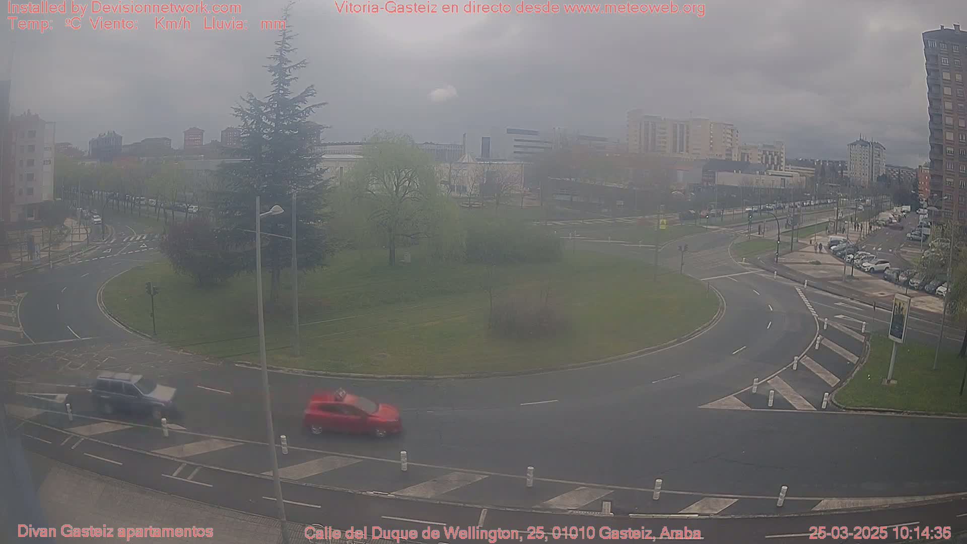 A hazy, overcast day shows a roundabout with a few cars and a grassy center, surrounded by buildings and parked cars.