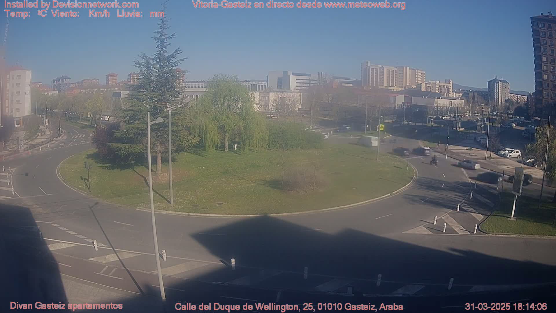 A sunny day reveals a traffic circle surrounded by buildings, green spaces, and several cars.