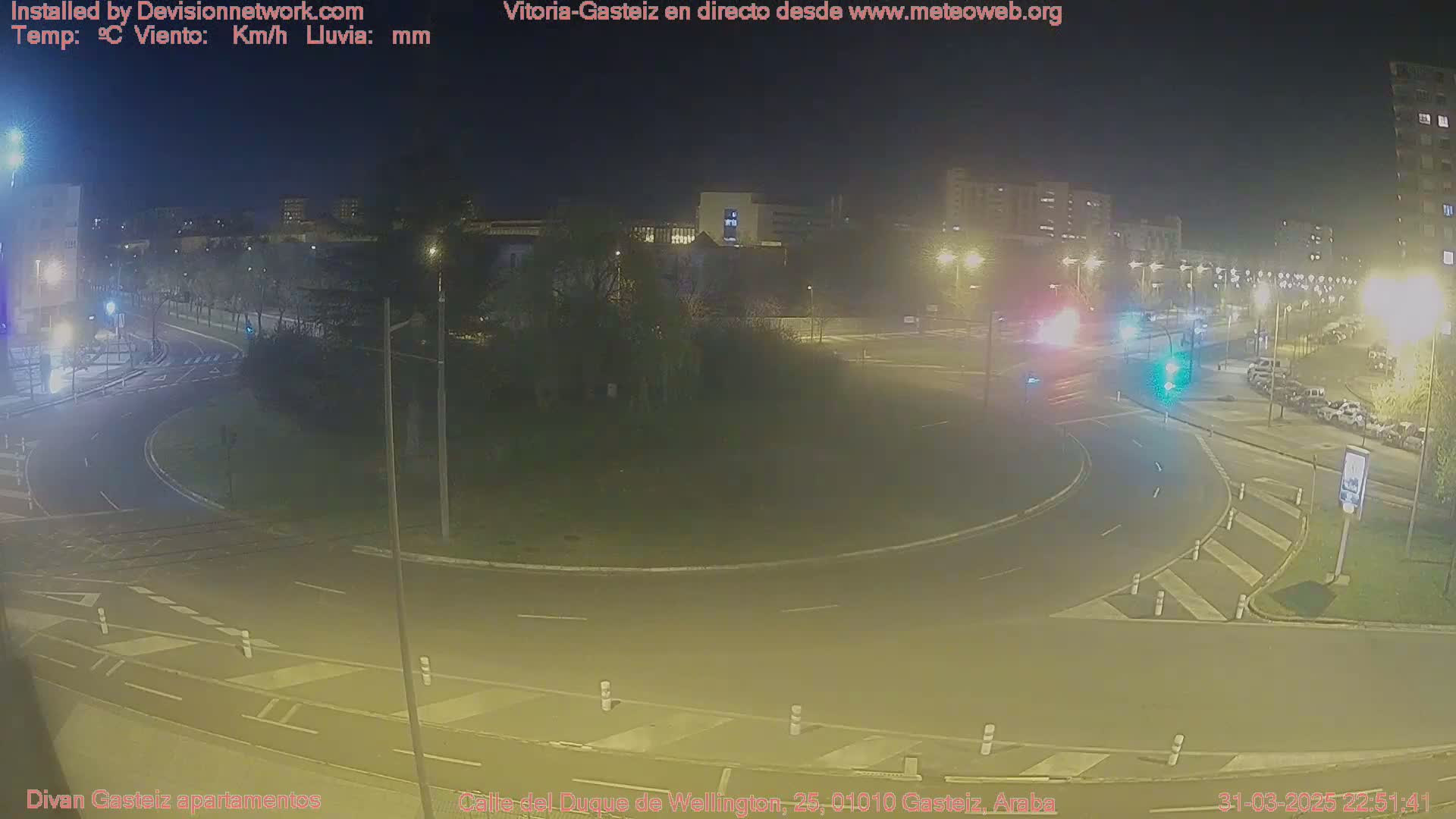 A nighttime view of a traffic circle with cars and buildings in the background under clear skies.