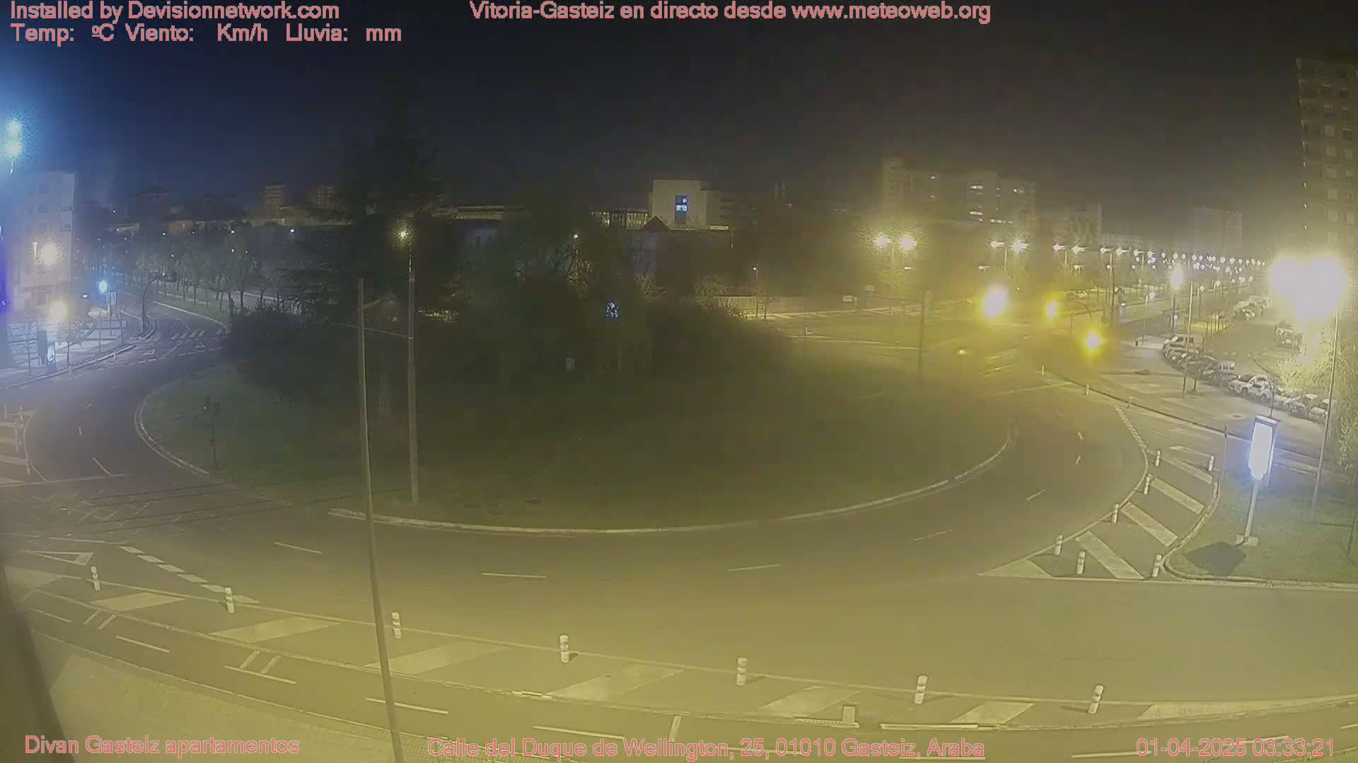 A nighttime view of a roundabout with cars and streetlights, surrounded by buildings and trees under a clear sky.