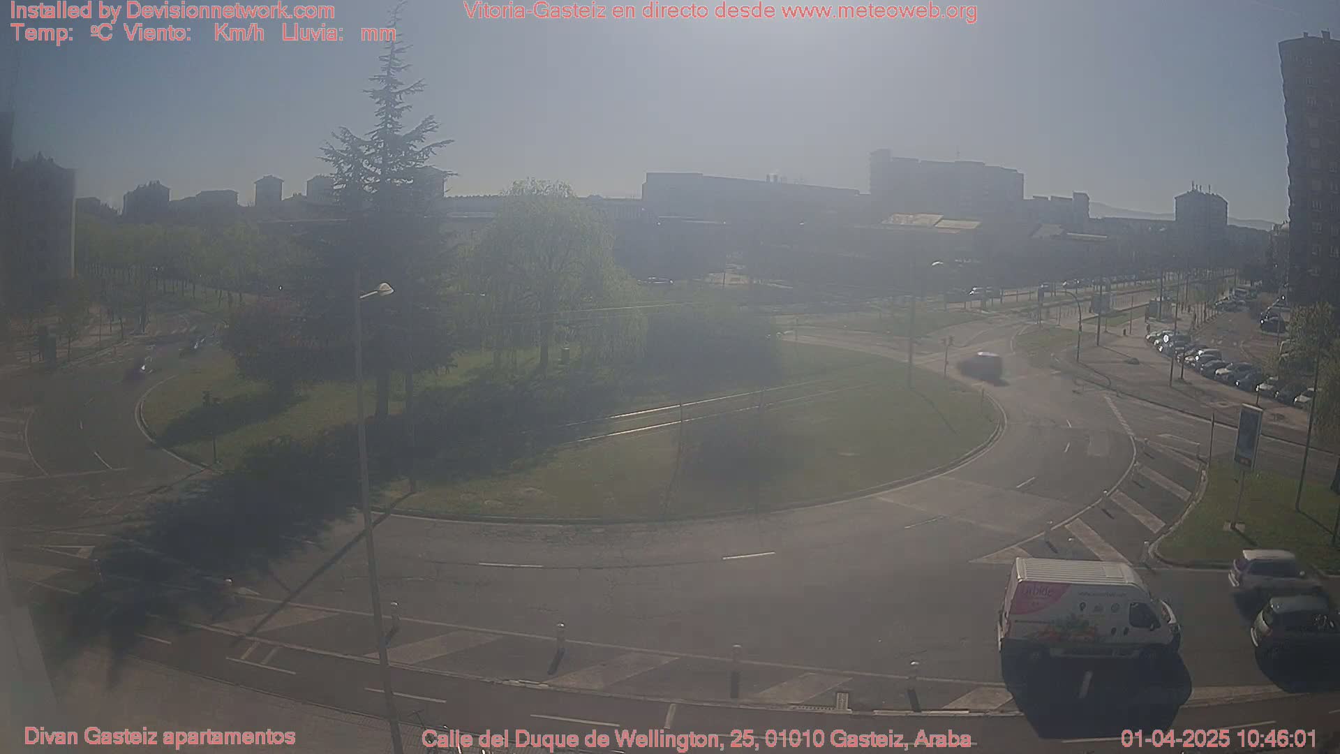 A hazy sunny day shows a roundabout with light traffic and surrounding buildings, greenery, and parked cars.