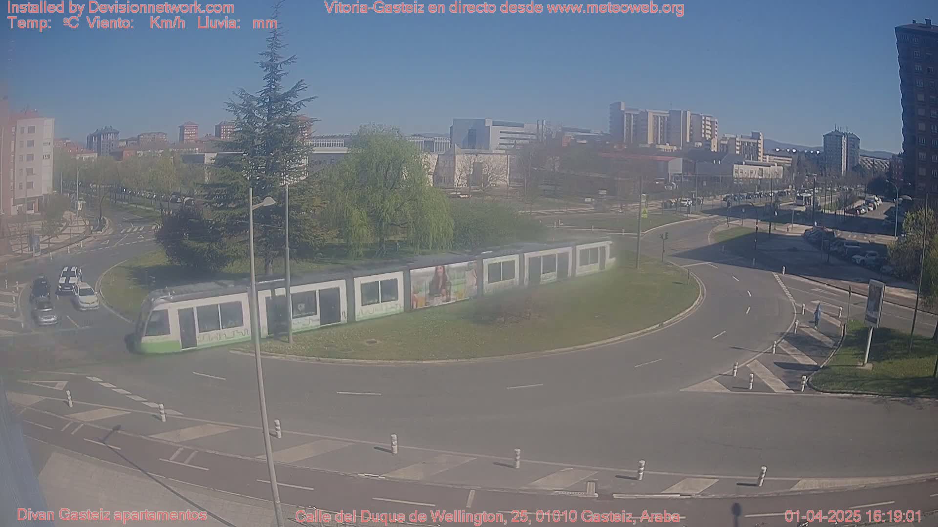A long, light-green tram sits in a roundabout in a city on a sunny day, with several cars visible on the roads around it and buildings in the background.