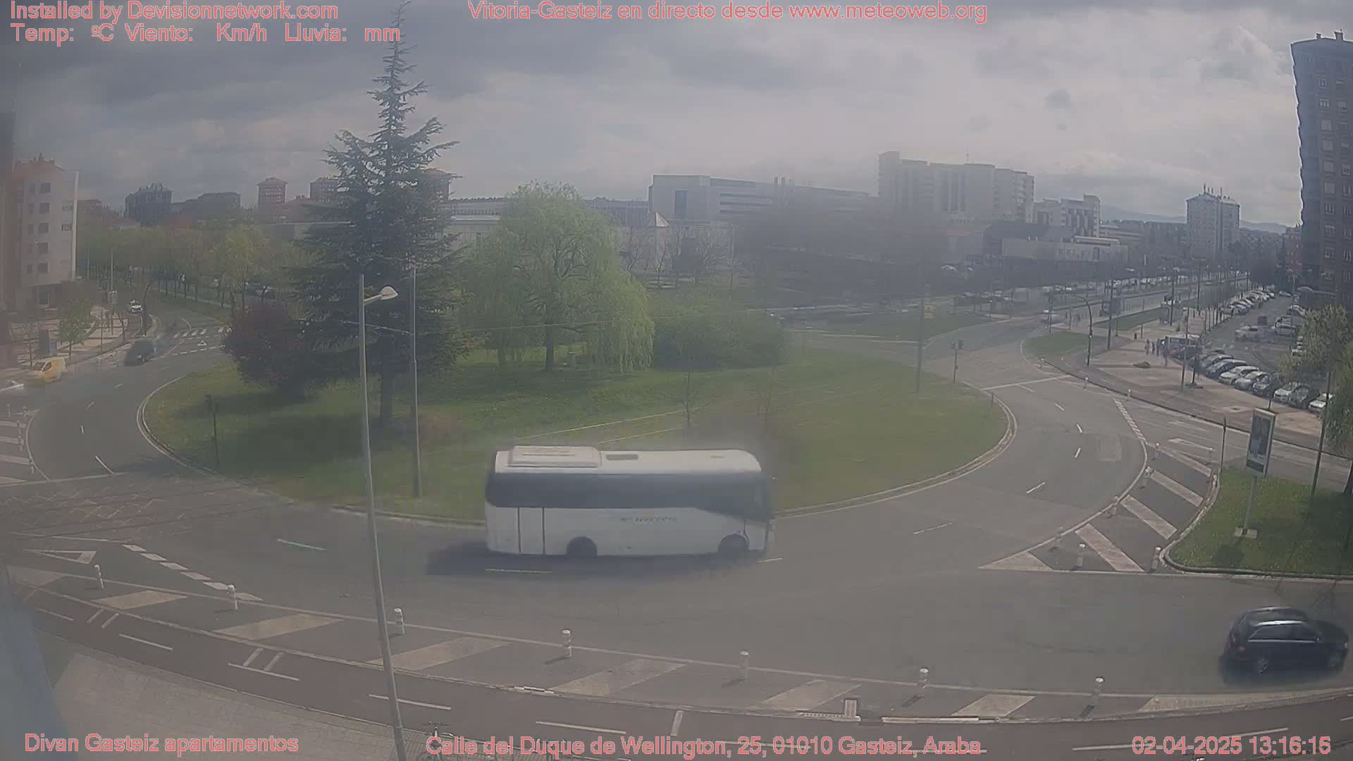 A hazy, overcast day shows a roundabout with a white bus and a dark car, surrounded by buildings and greenery.