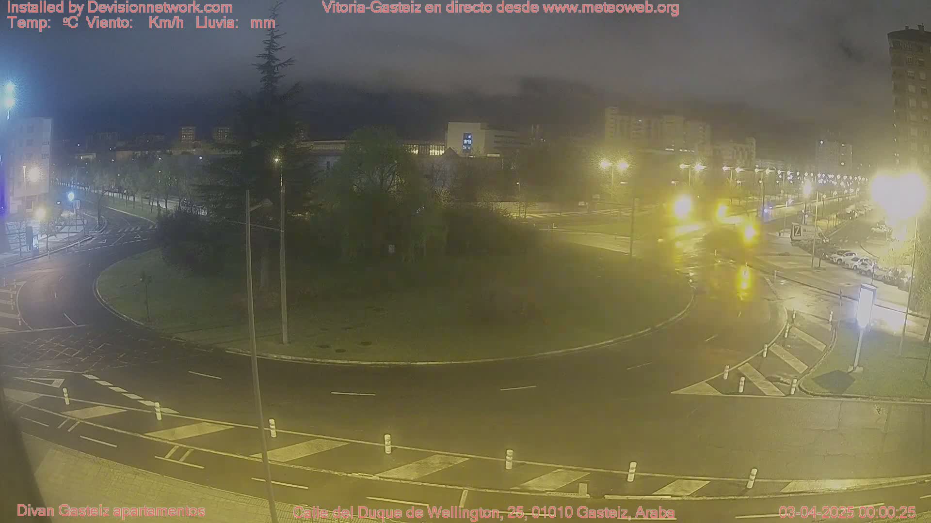 A nighttime view of a wet roundabout and surrounding streets, illuminated by streetlights, with buildings visible in the background.