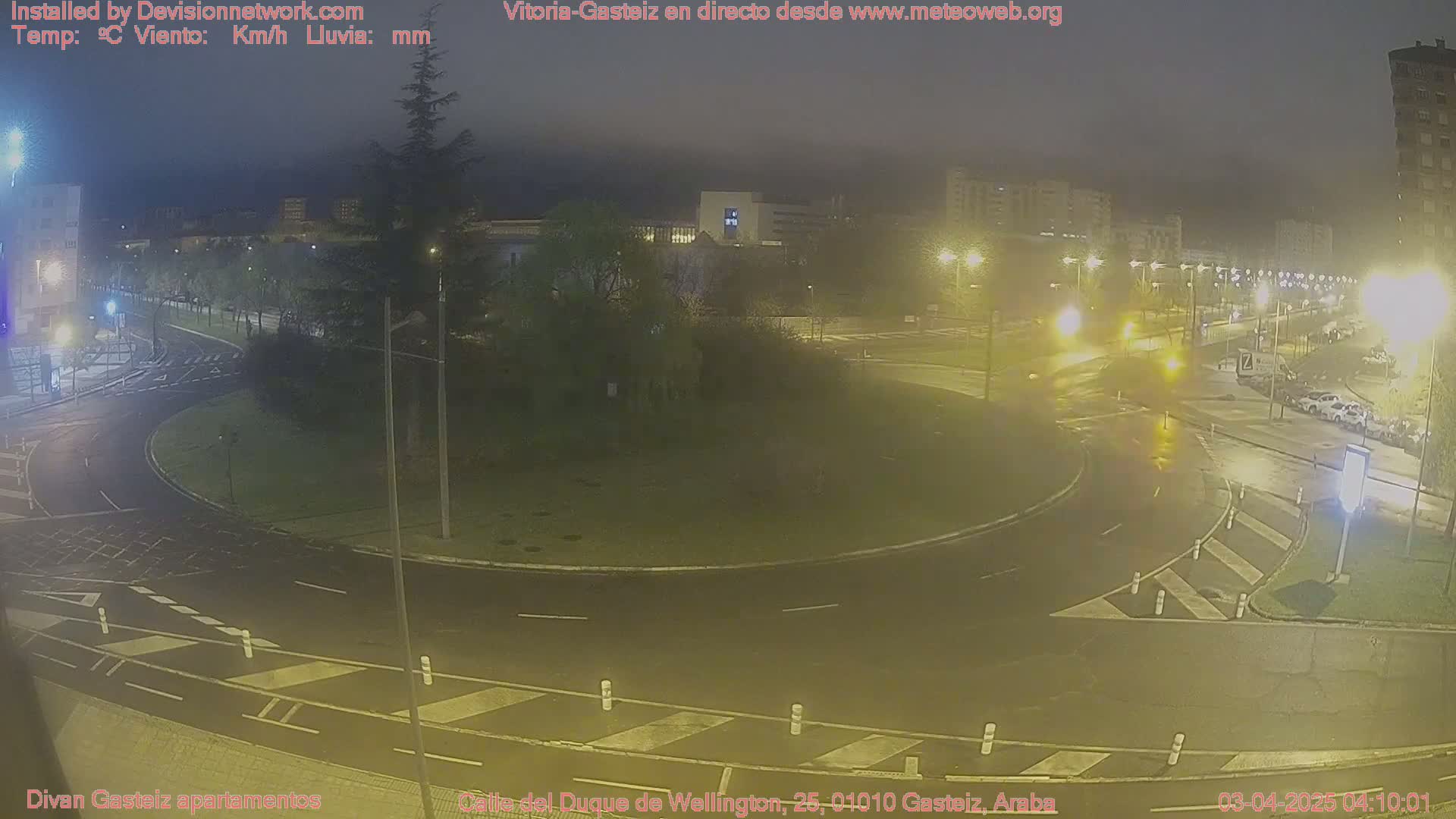 A rain-slicked roundabout at night, illuminated by streetlights, with several vehicles visible and buildings in the background.