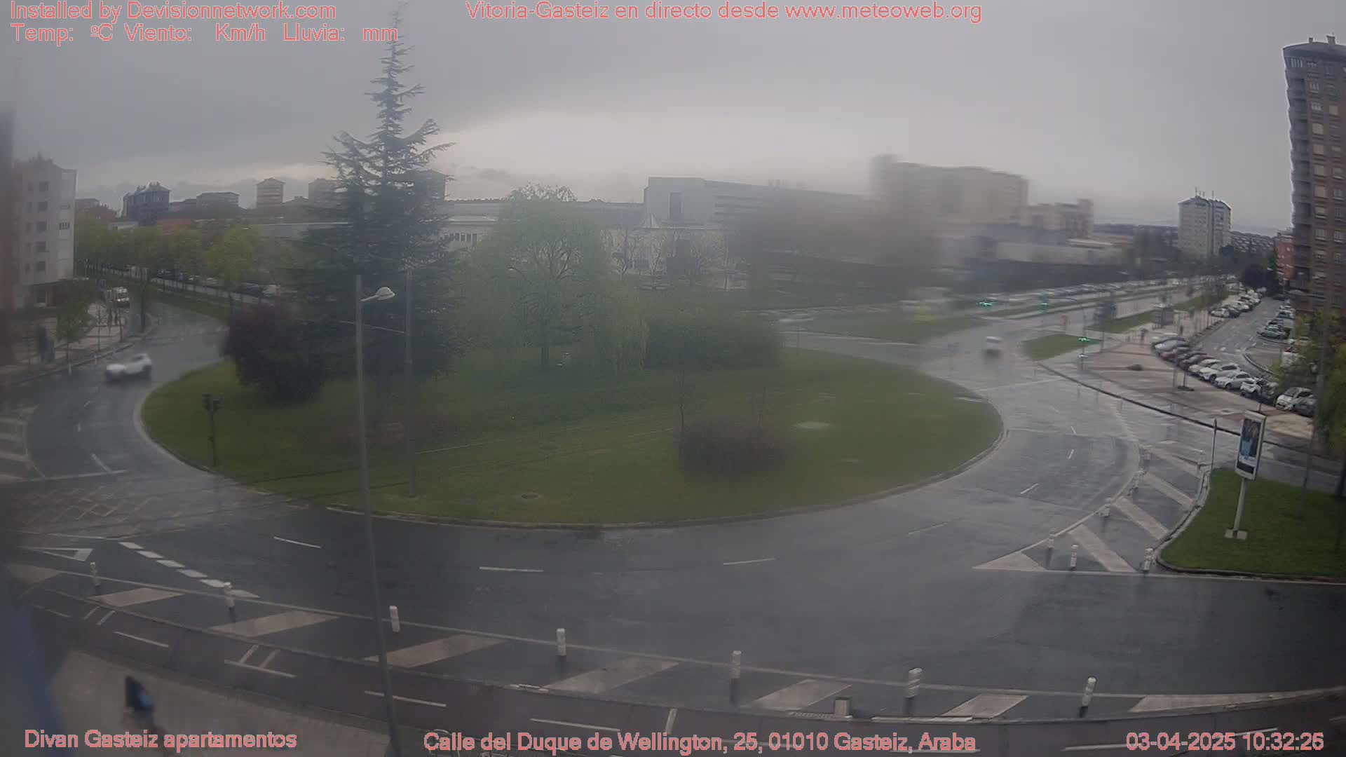 A wet, overcast scene shows a roundabout in a city with several cars driving around it, with buildings and trees visible in the background.