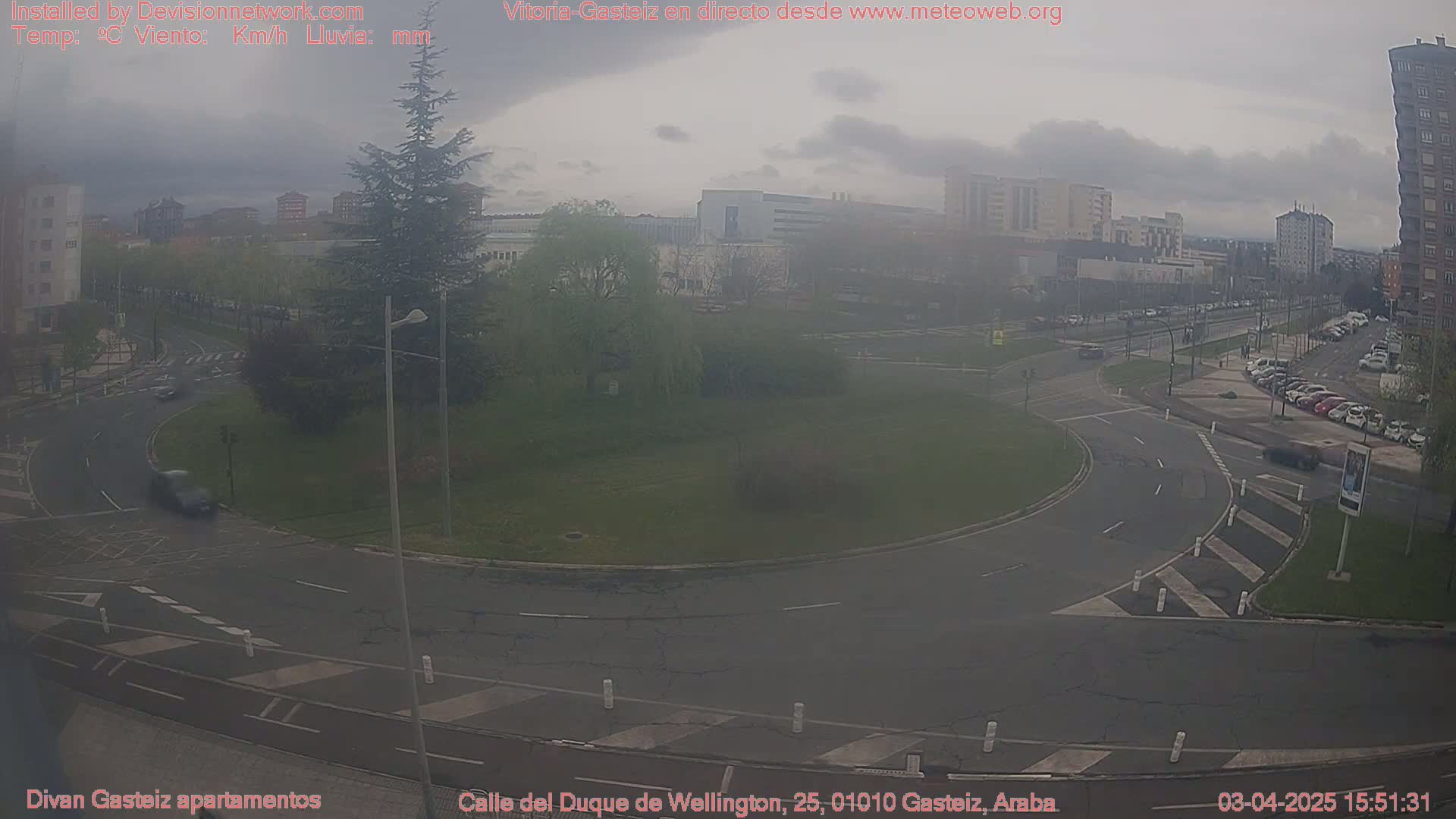 A hazy overcast day reveals a roundabout surrounded by buildings and parked cars, with a few vehicles visible on the road.