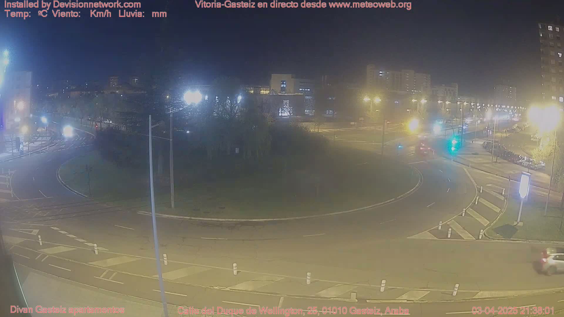 A nighttime view of a roundabout in a city, with cars driving around it and buildings visible in the background under clear skies.