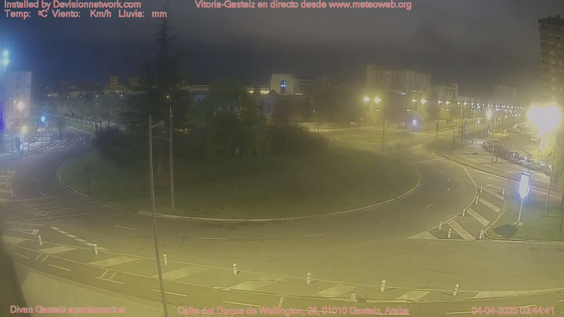 A nighttime, dimly lit view of a roundabout and surrounding streets in a city, with some parked cars visible.