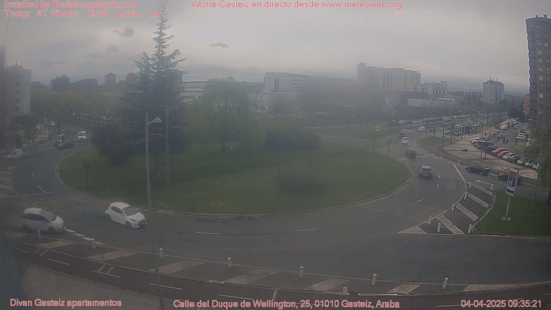 A hazy, overcast day reveals a roundabout with several cars and a tram in the distance, surrounded by buildings and greenery.