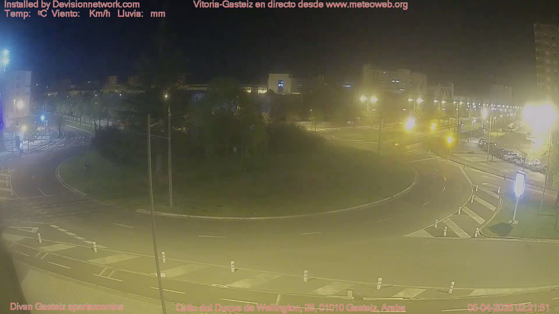 A nighttime view of a roundabout and surrounding roads, illuminated by streetlights, with parked cars visible in a nearby lot.