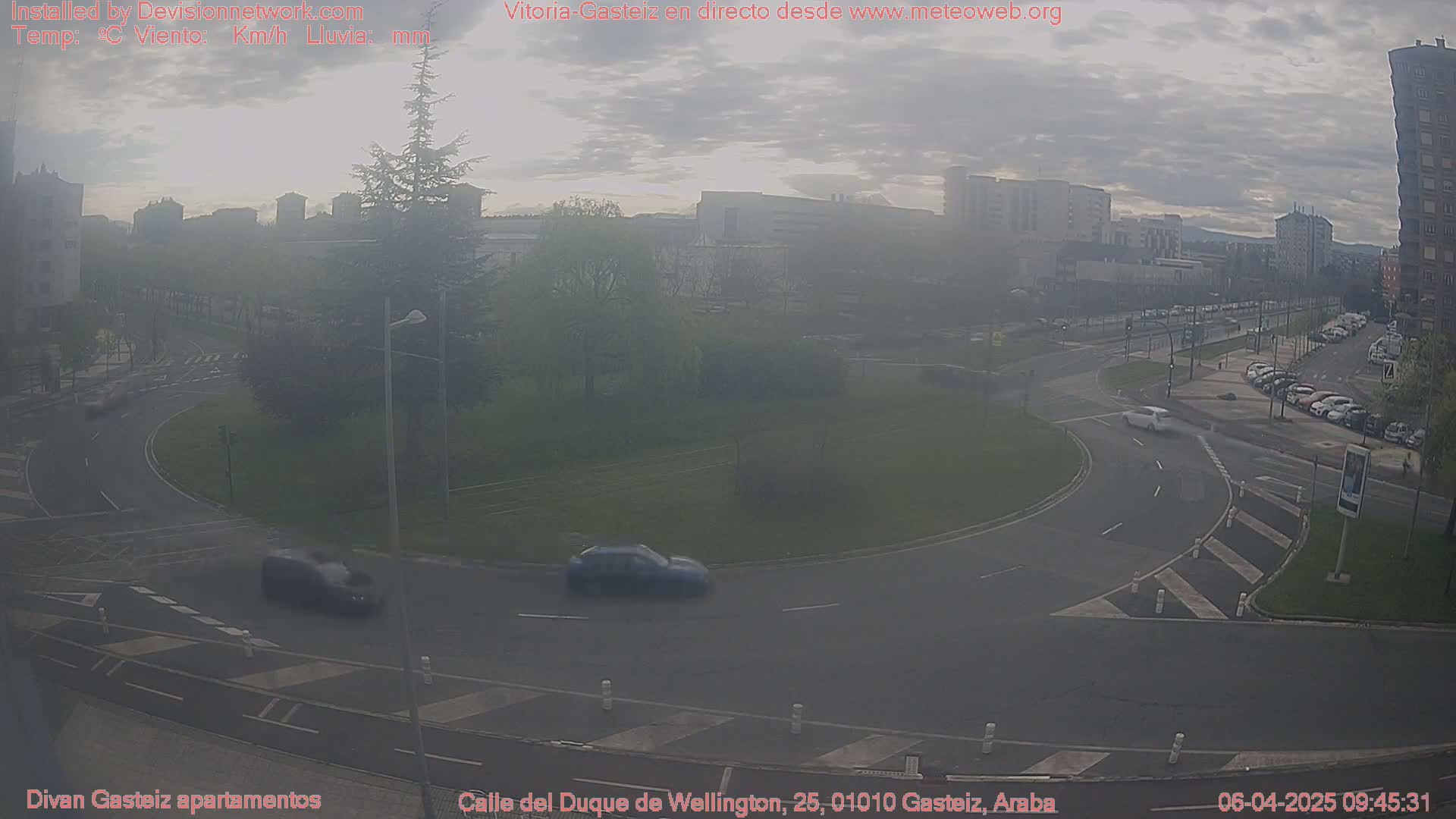 A hazy, overcast day reveals a traffic circle in an urban area with several cars visible, surrounded by buildings and green spaces.