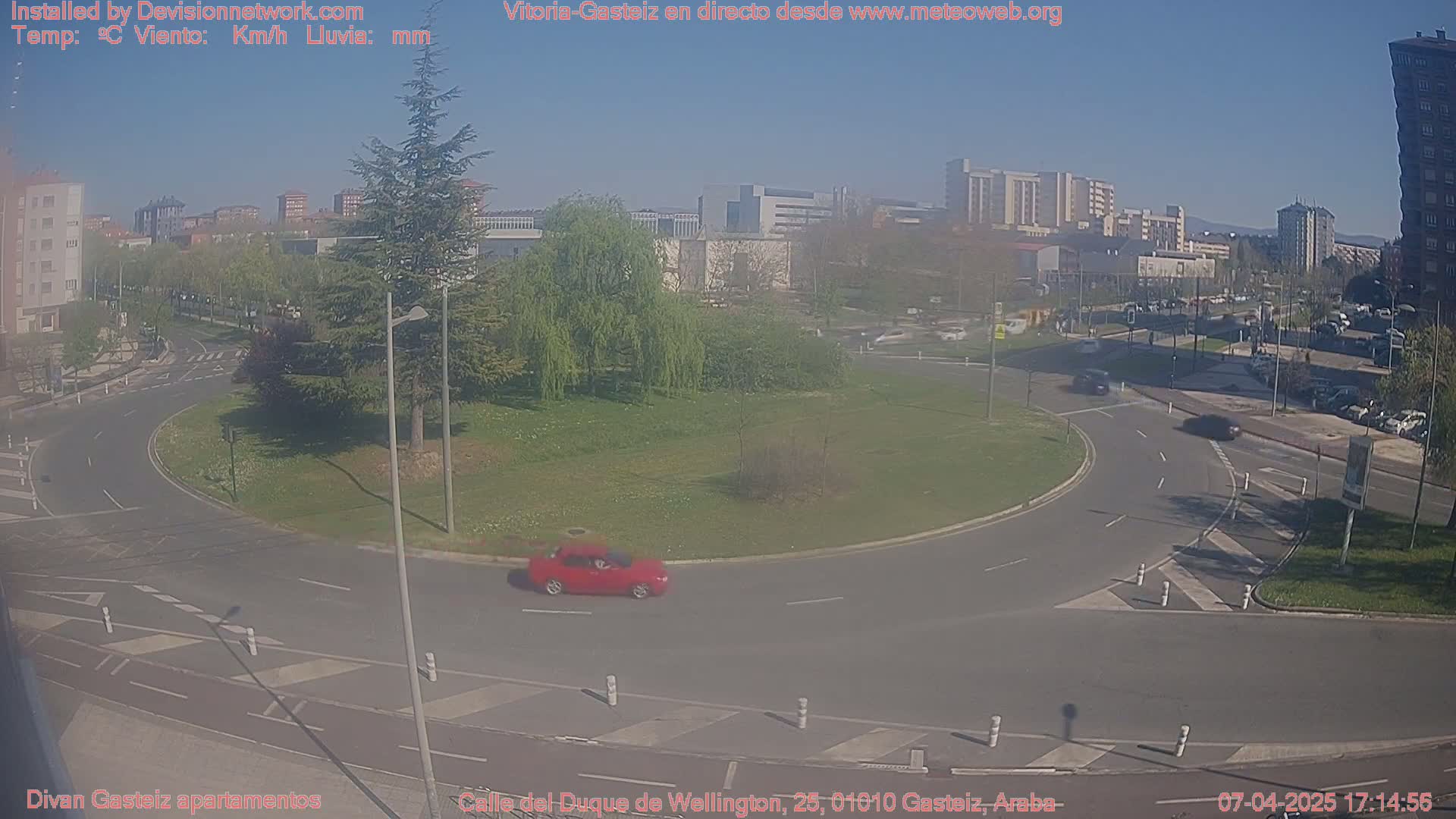 A sunny day shows a red car driving around a traffic circle in a city, with several buildings and trees visible in the background.