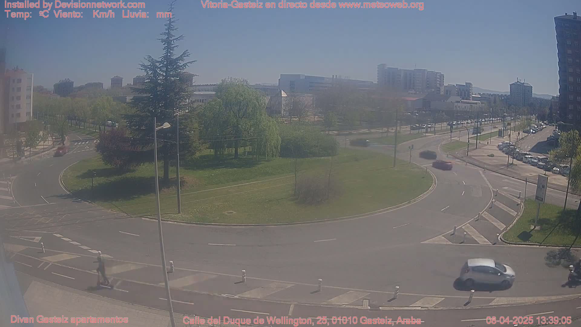 A sunny day shows a traffic circle in a city with several cars, a person on a scooter, and buildings in the background.