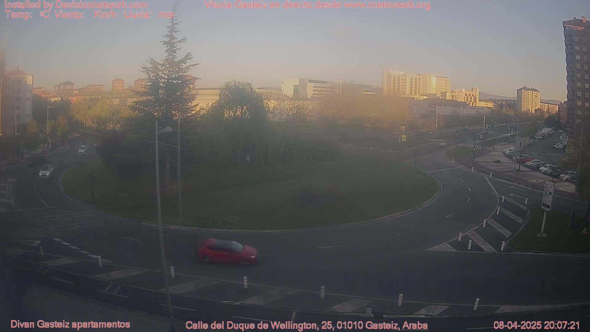 A hazy sunset illuminates a roundabout in a city, with several cars visible on the road and parked in a lot, and buildings in the background.
