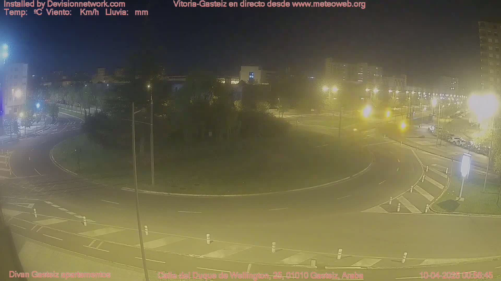 A nighttime view of a traffic circle, illuminated by streetlights, with cars visible on the roads and parked along the curb.