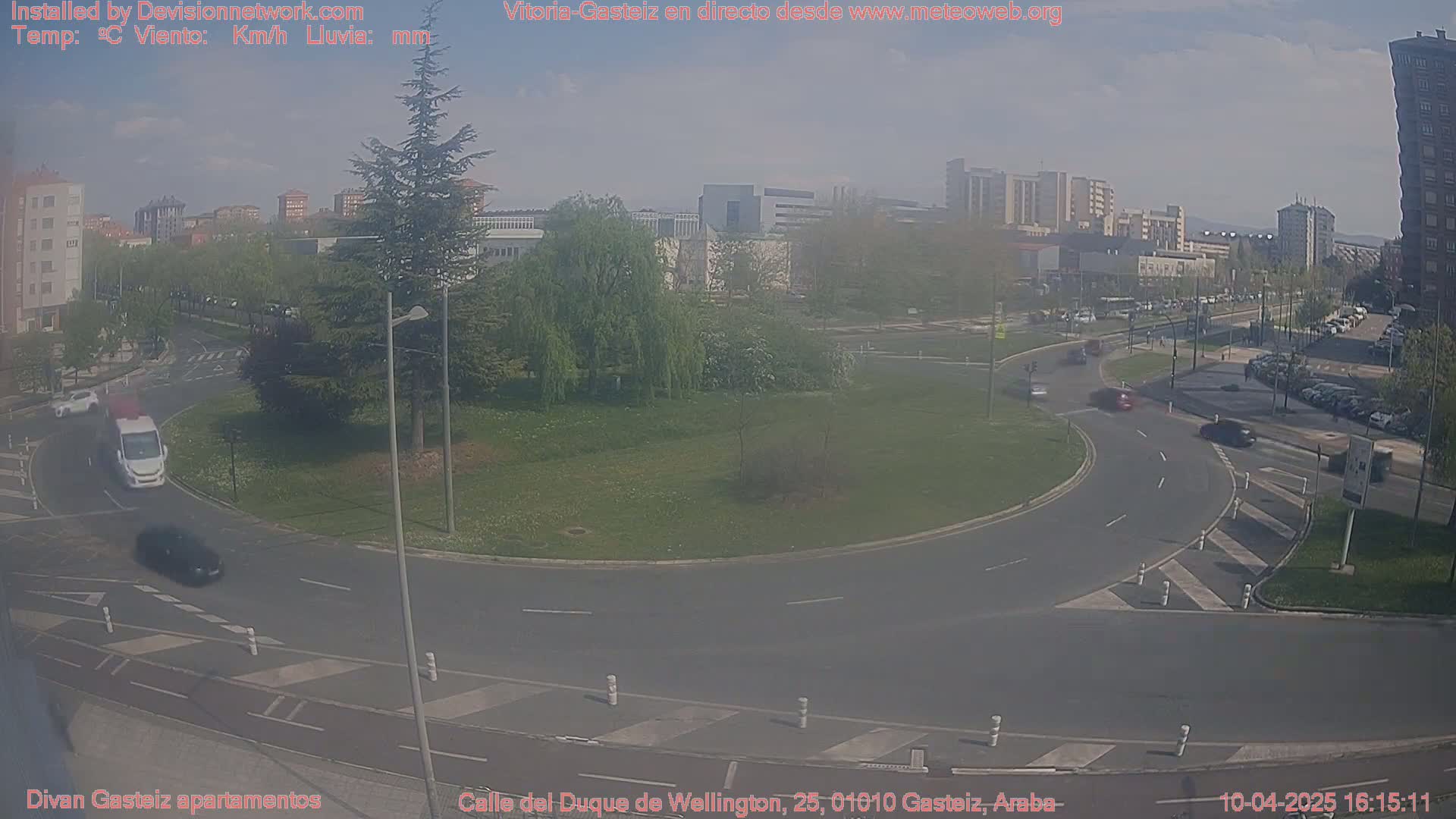 A partly cloudy day shows a roundabout with several cars driving on roads near apartment buildings and trees.