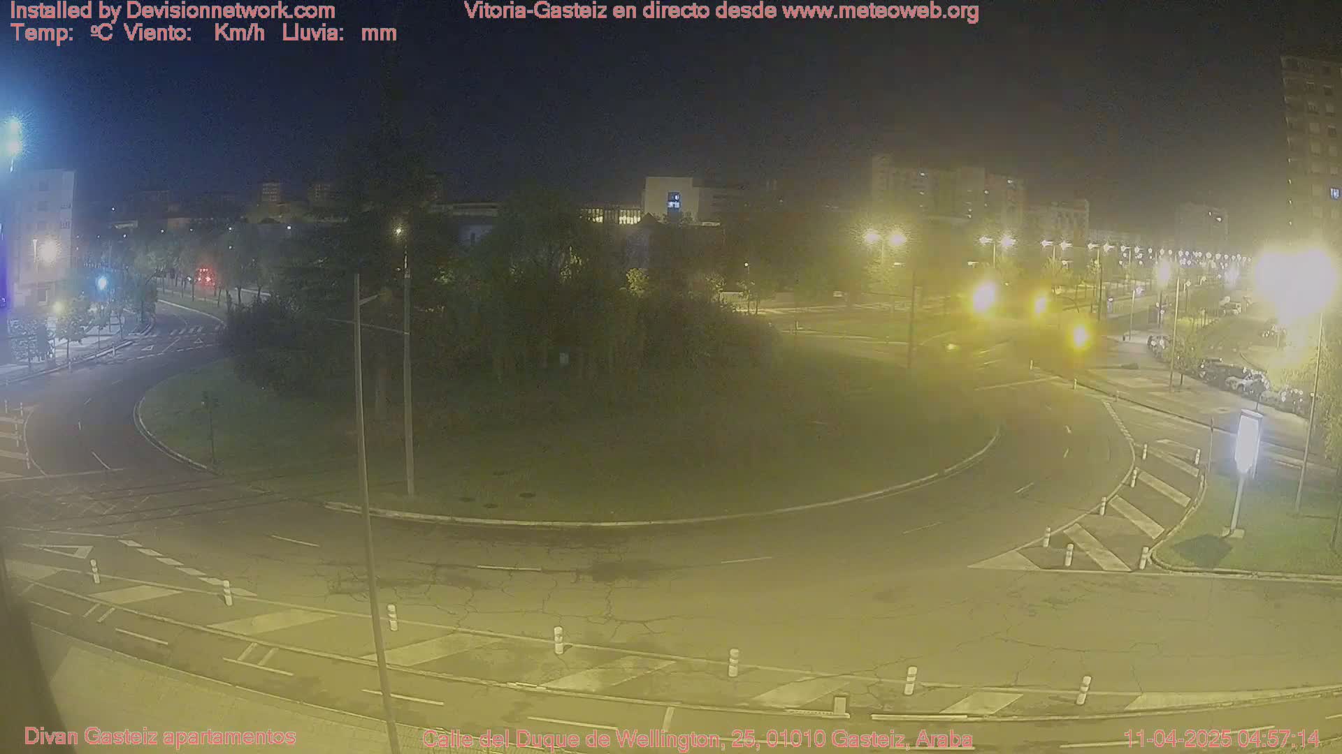 A nighttime, overhead view of a roundabout surrounded by roads, buildings, and trees under dark, well-lit conditions.