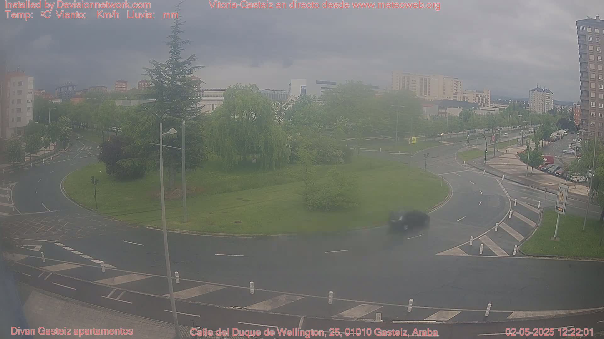A roundabout in a city, under an overcast sky, with cars driving on the roads and green spaces around it.