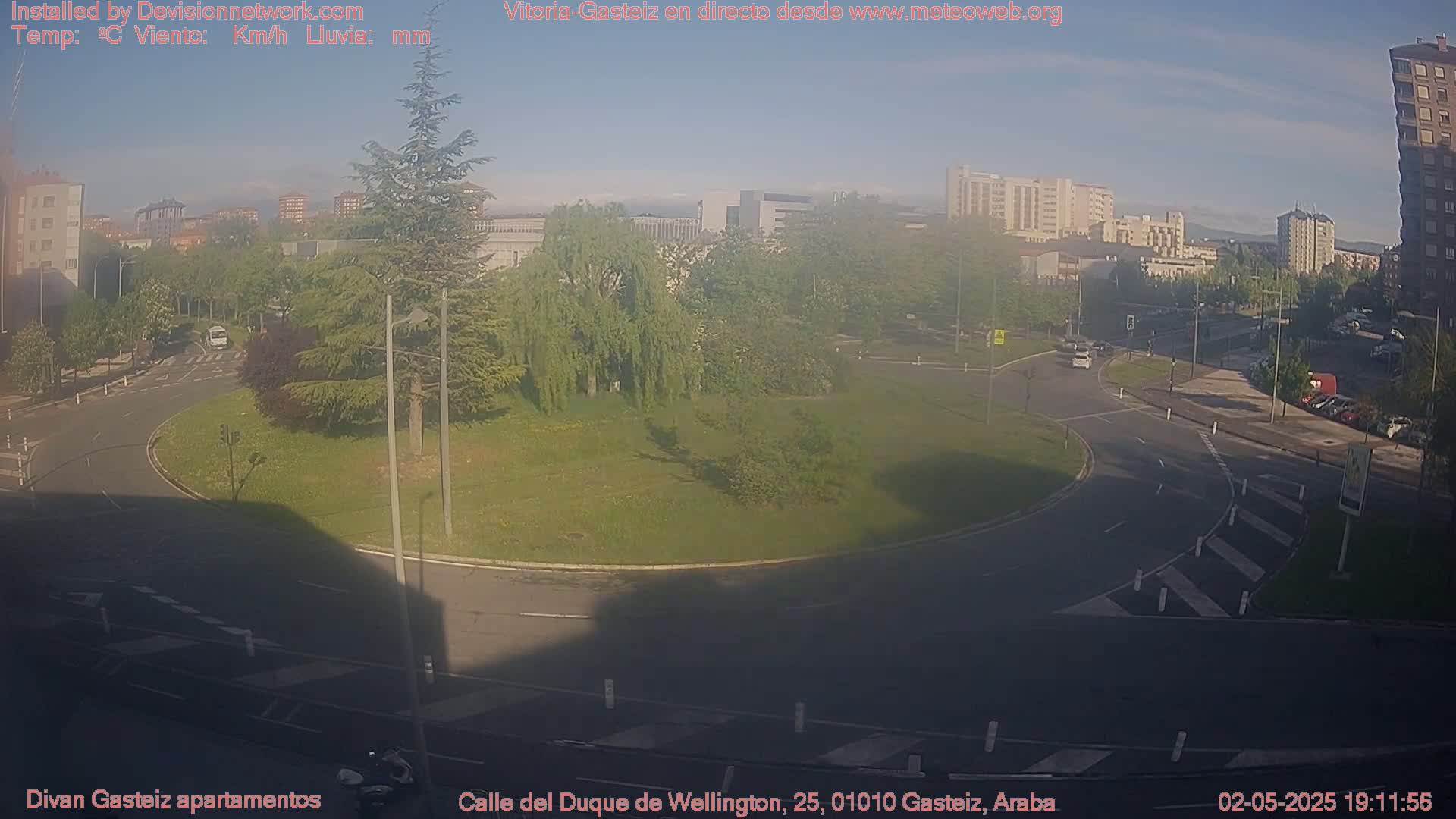 A sunny view of a roundabout surrounded by grassy areas and buildings, with a few cars visible on the road.