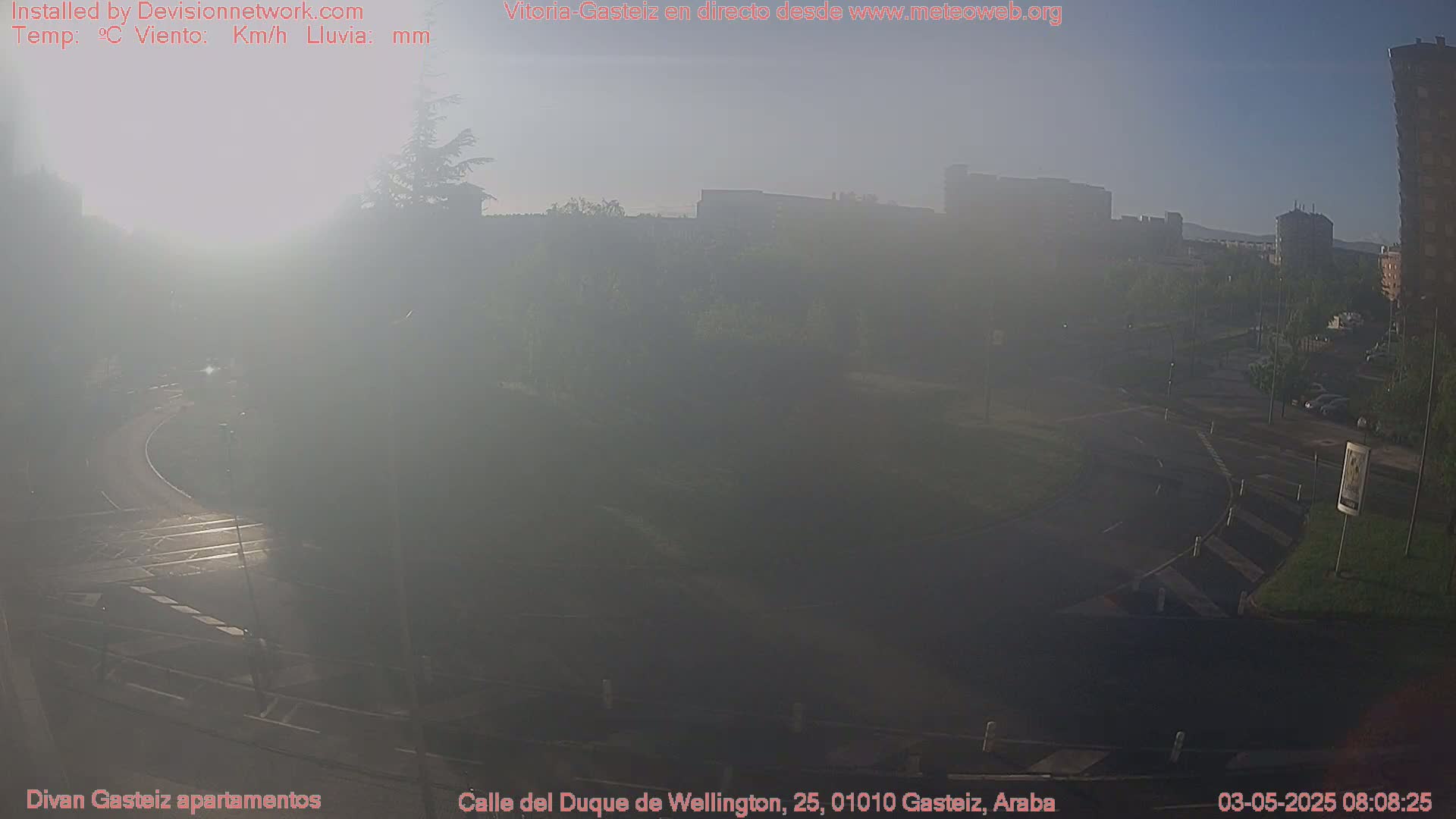 A hazy, sunlit view of a roadway intersection with buildings and trees in the background.
