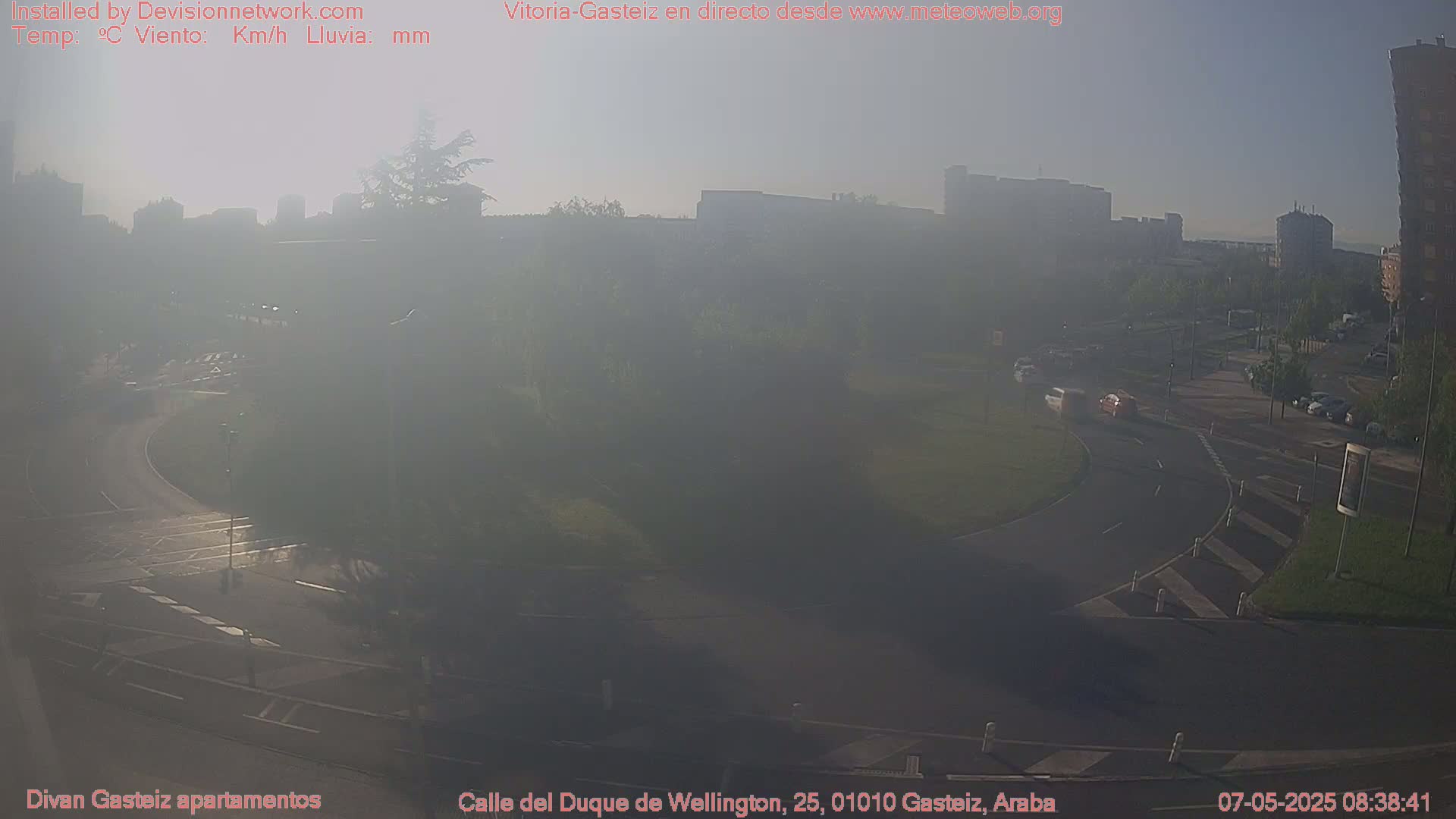 A hazy, sunrise view of a roadway intersection with sparse traffic in front of a city skyline.