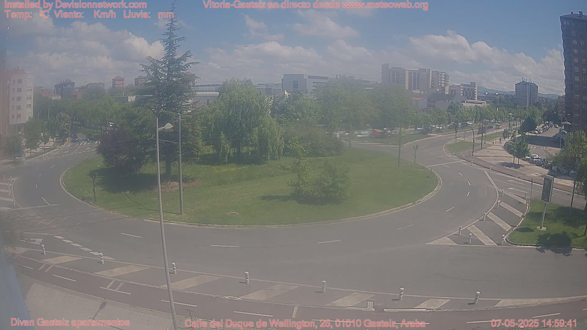 A mostly sunny day reveals a roundabout intersection surrounded by green landscaping and city buildings in the background.