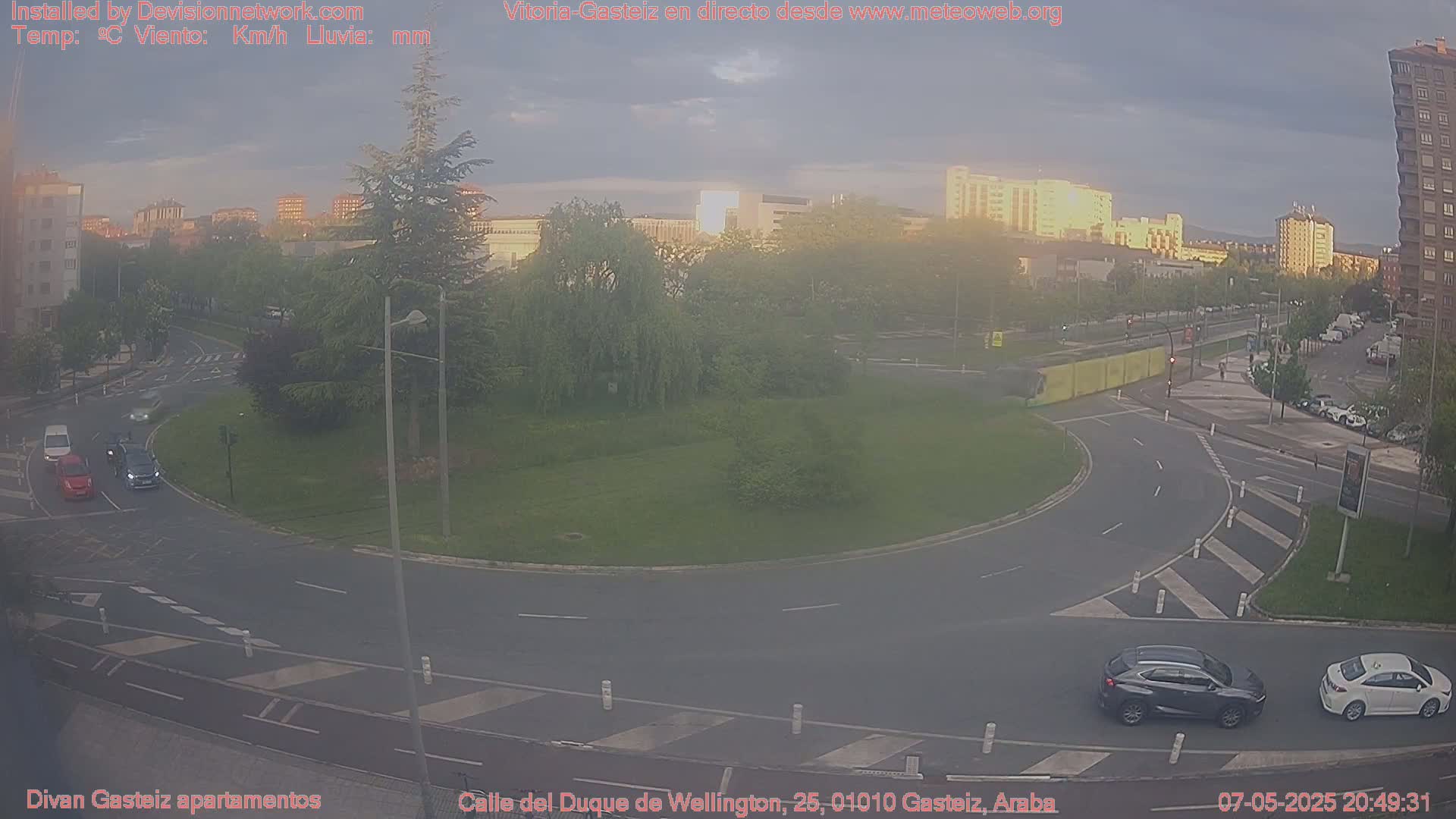A mostly clear evening shows a traffic circle surrounded by green spaces and buildings, with several cars visible.