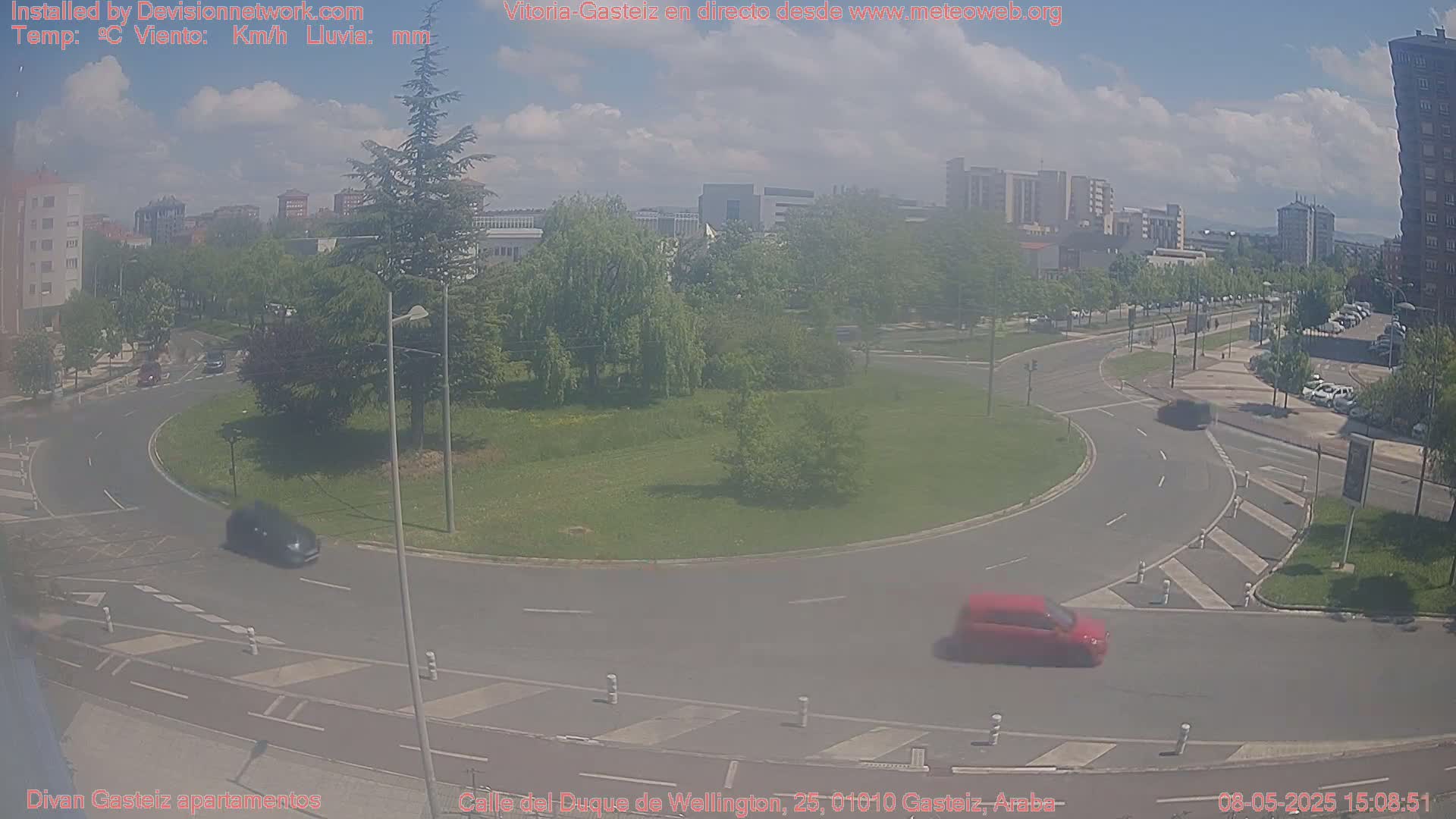 A partly cloudy day shows a roundabout with cars driving on surrounding roads and buildings in the background.