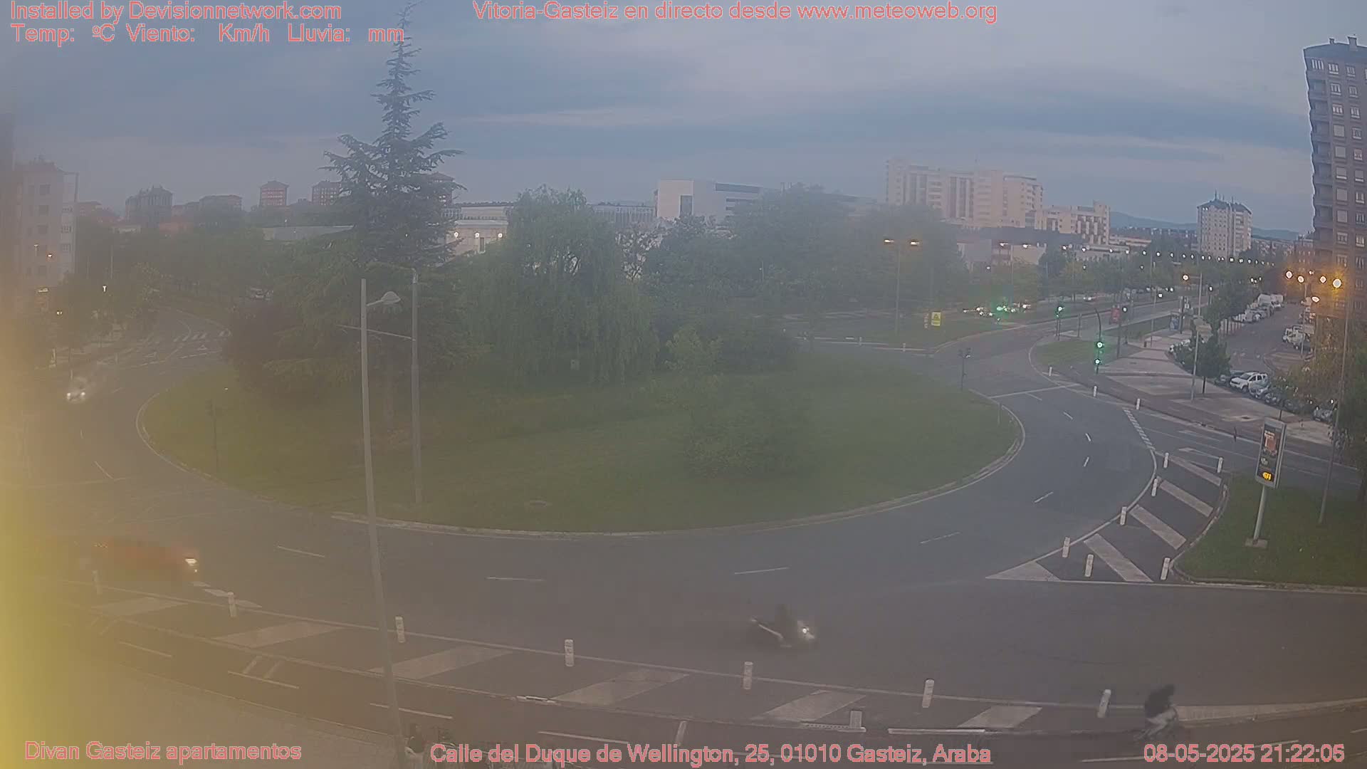 A hazy, overcast evening scene shows a roundabout surrounded by buildings and trees, with several vehicles visible on the roads.