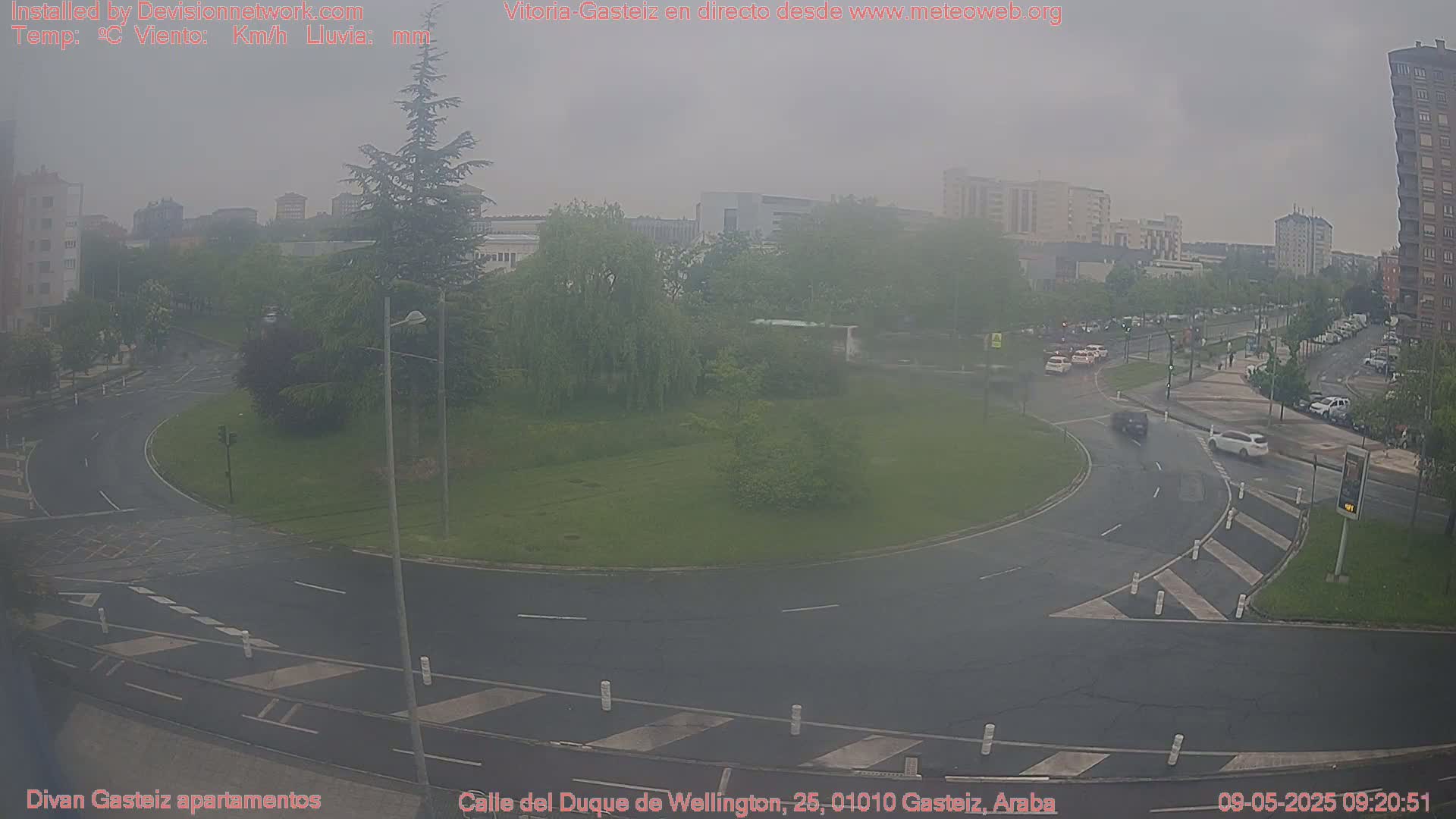 A hazy, overcast day reveals a roundabout surrounded by green space and buildings, with several cars visible on the road.