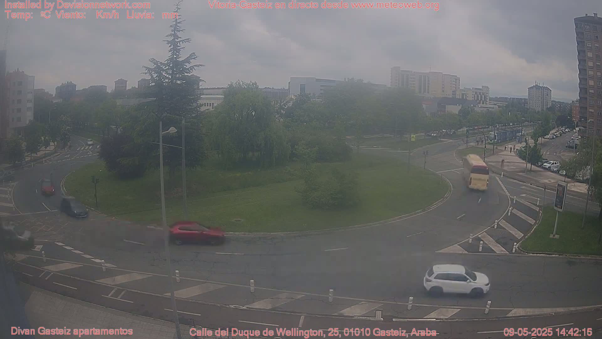 A hazy overcast day reveals a roundabout with several cars and a bus navigating its lanes, surrounded by city buildings and greenery.