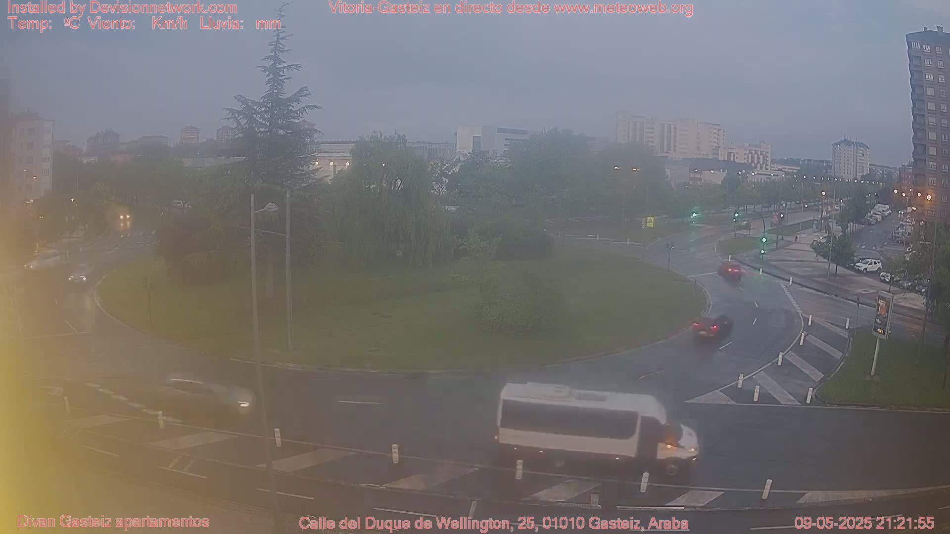 A wet roundabout with several cars and a van driving through it, surrounded by greenery and buildings under an overcast sky.
