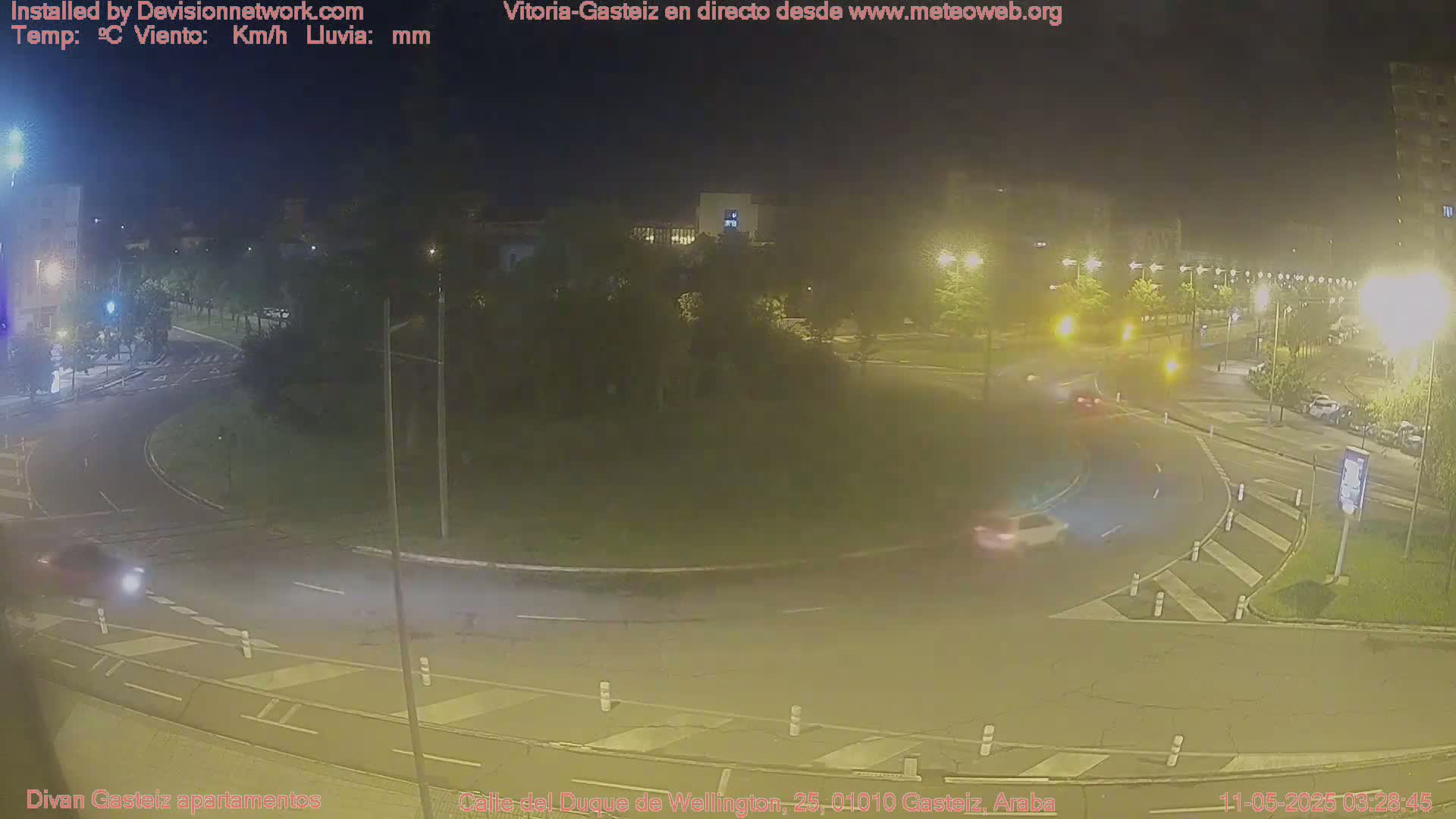 A nighttime, slightly hazy view of a roundabout with several cars driving around it and streetlights illuminating the scene.