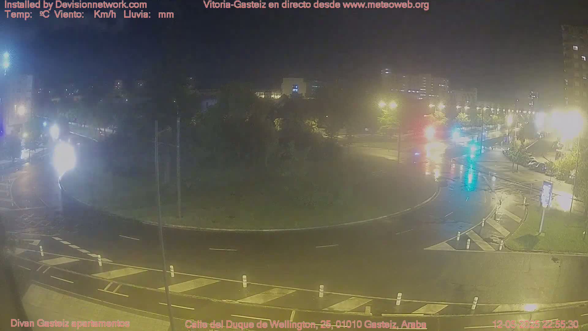 A wet, nighttime view of a roundabout with several cars, surrounded by buildings and streetlights.