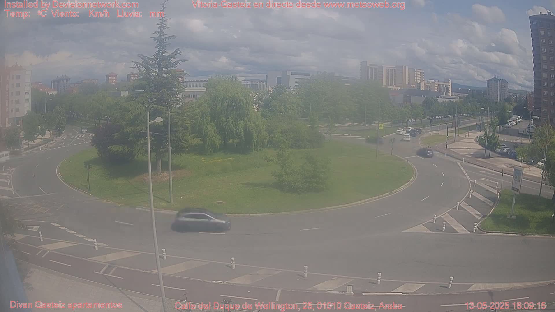 A hazy view of a roundabout with cars driving around it, surrounded by green grass, trees, and buildings under a partly cloudy sky.