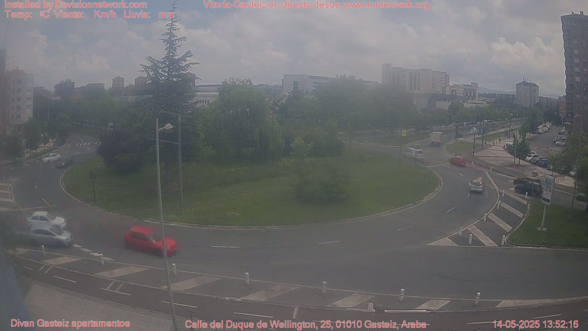 A hazy view of a roundabout with several cars driving around it, surrounded by buildings and green spaces.