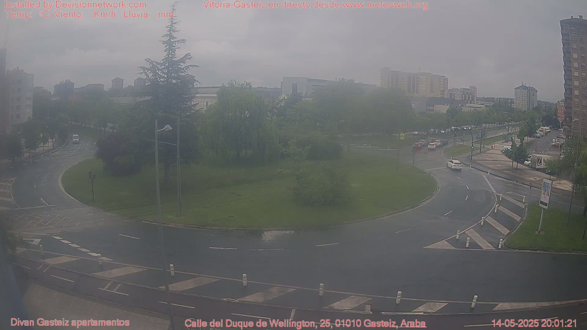 A rainy, overcast view of a traffic circle in a city, surrounded by buildings and trees.