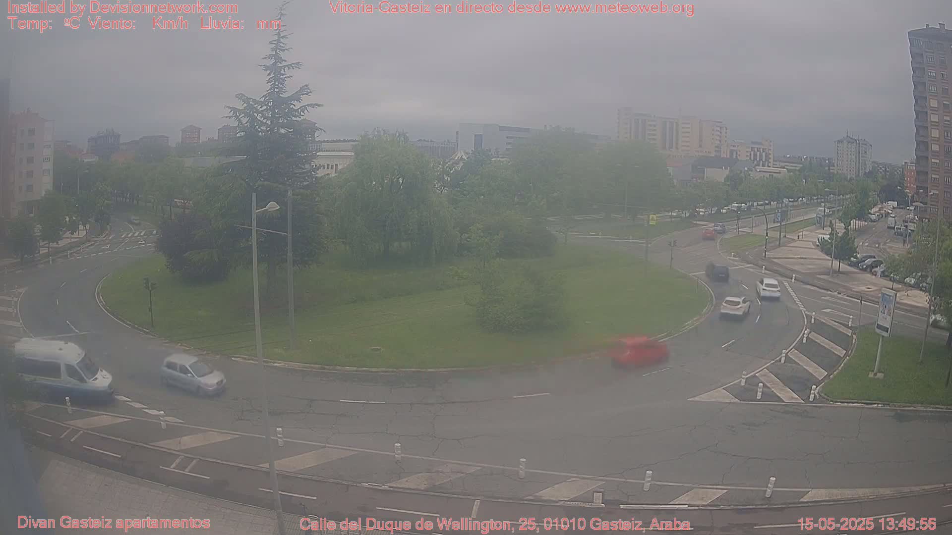 A hazy, overcast view of a roundabout with several cars and a van, surrounded by buildings and trees.