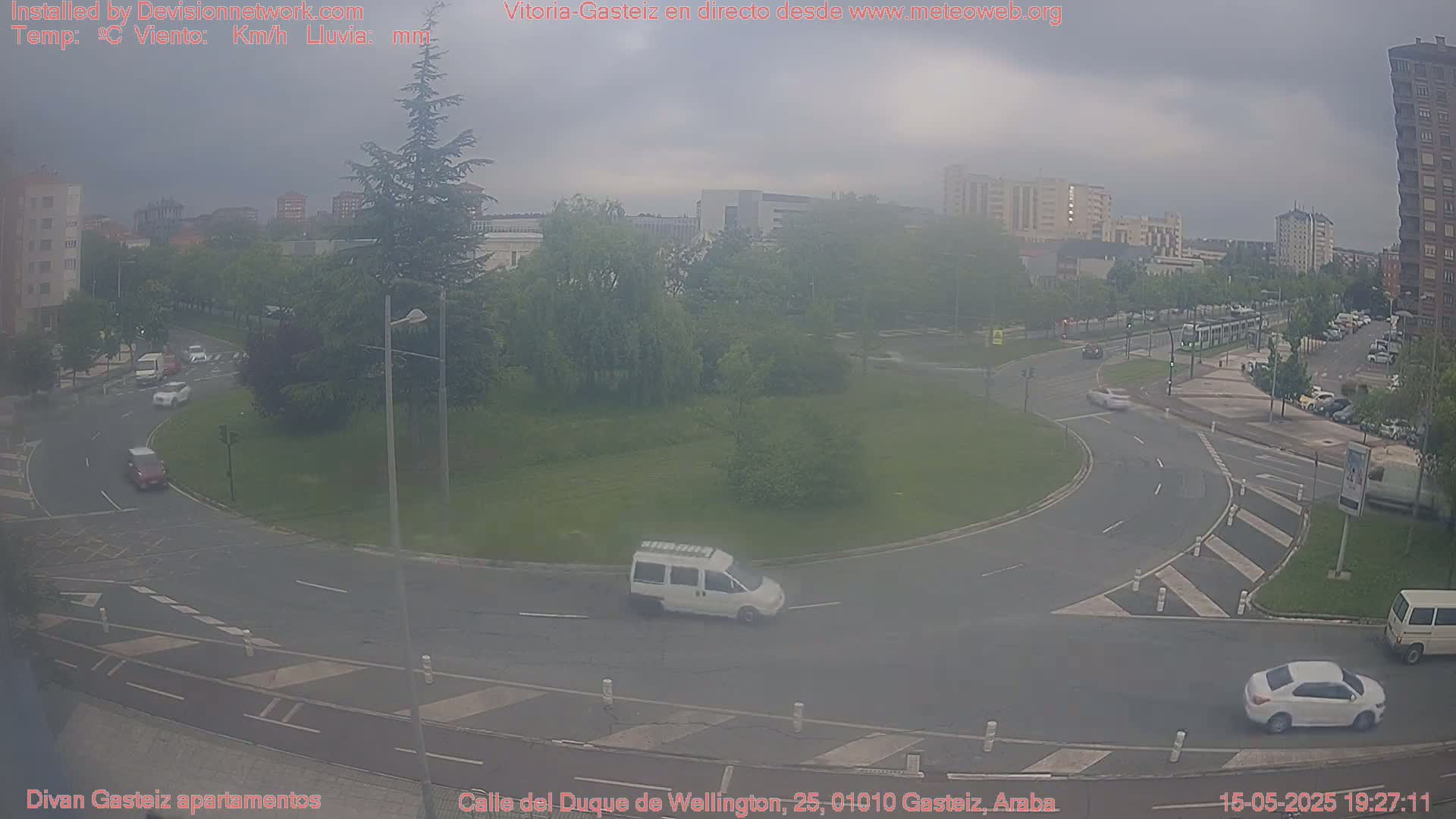 A hazy overcast day reveals a roundabout with several cars and a van, surrounded by green spaces and buildings in the background.