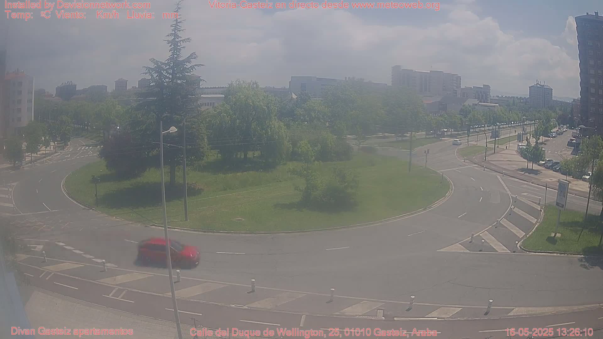 A hazy view of a traffic circle surrounded by green space and buildings under a partly cloudy sky.