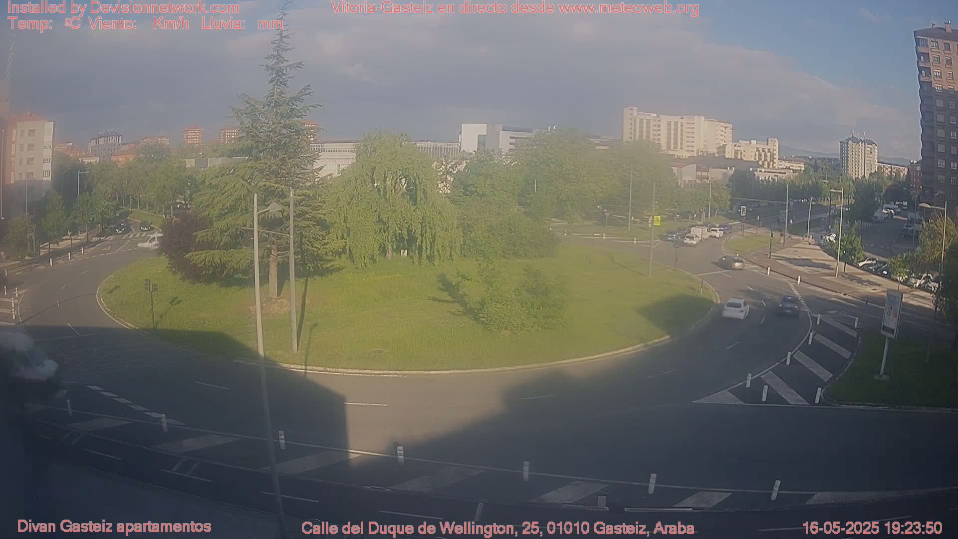 A mostly sunny day shows a roundabout in a city, with several cars driving around and through it, surrounded by buildings and trees.