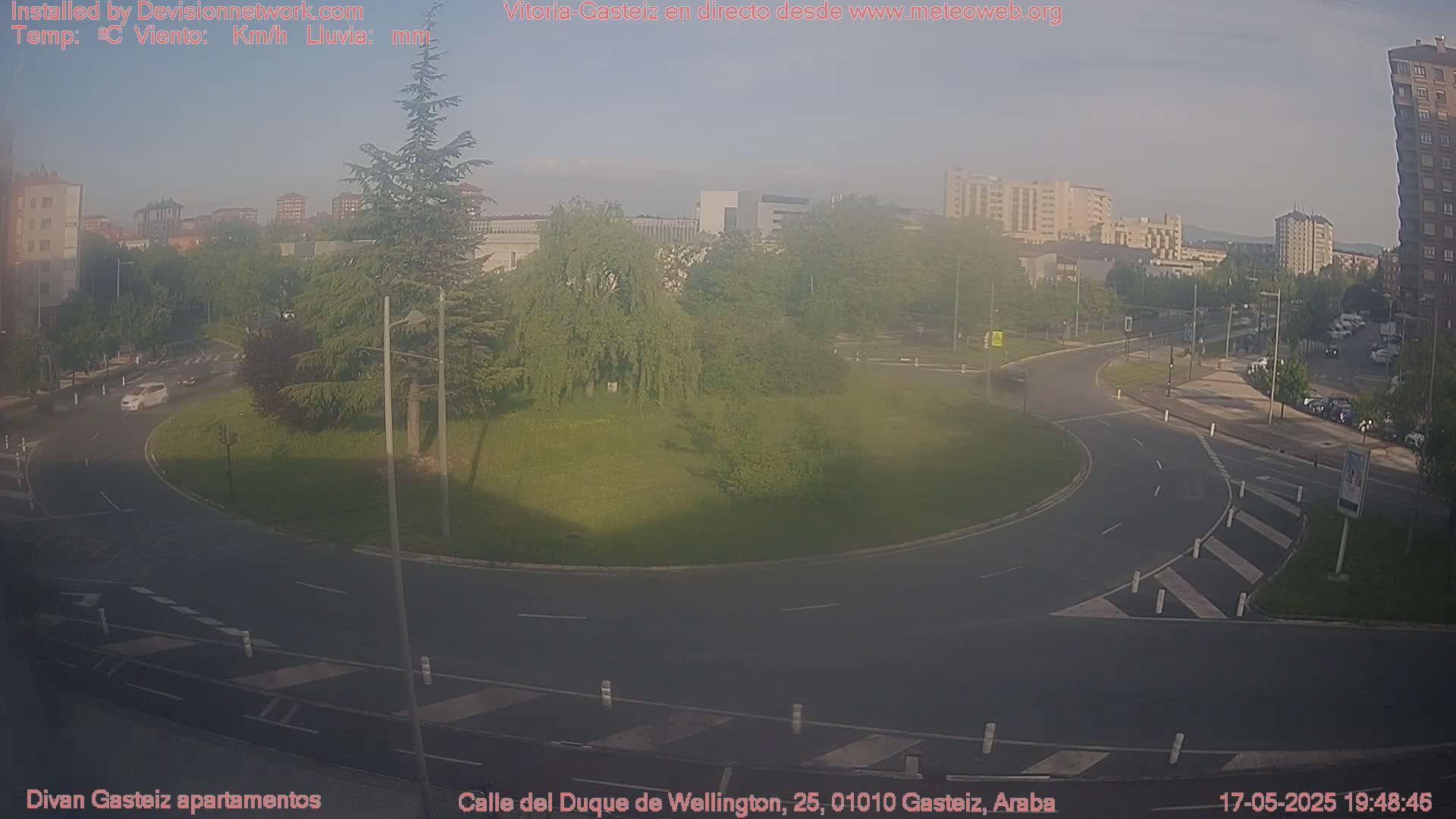 A hazy view of a roundabout with cars, surrounded by green grass, trees, and buildings under a mostly clear sky.