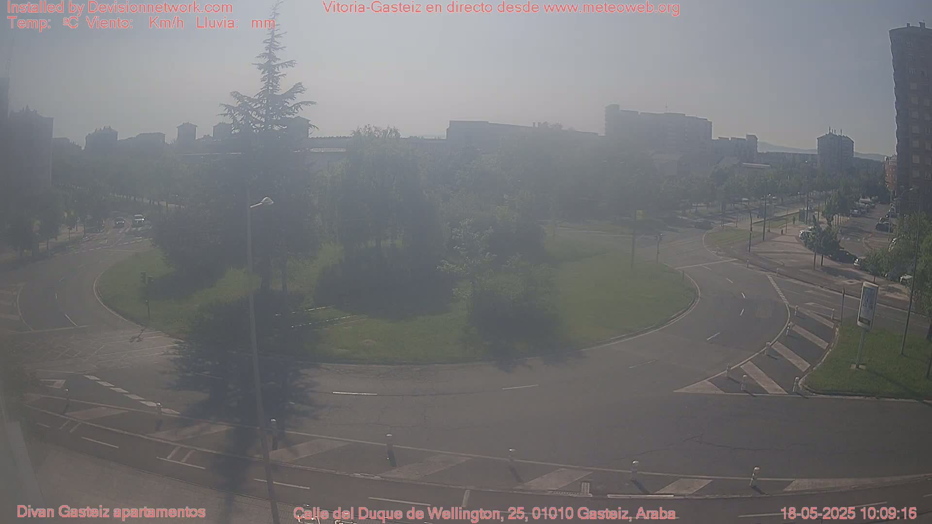 A hazy, sunny day shows a roundabout and surrounding roads with buildings and trees in the background.