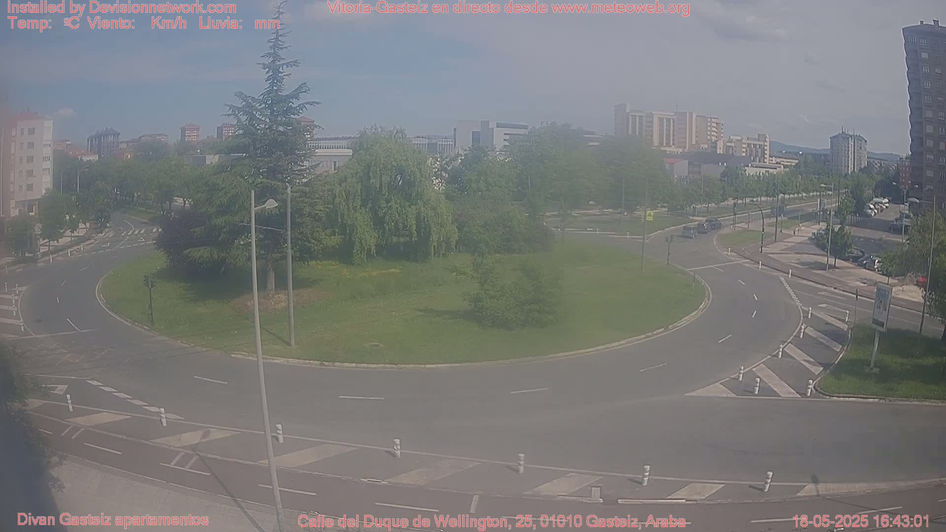 A hazy view of a roundabout in a city, surrounded by buildings, trees, and parked cars on a mostly sunny day.
