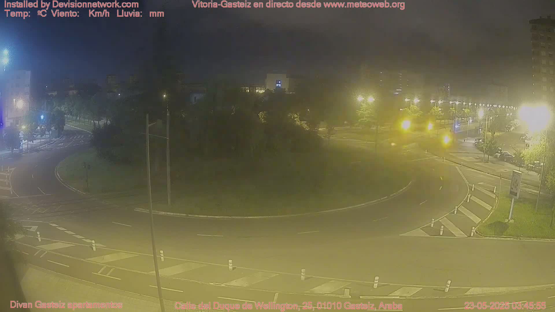 A nighttime view of a roundabout with streetlights illuminating a mostly empty roadway and surrounding greenery.