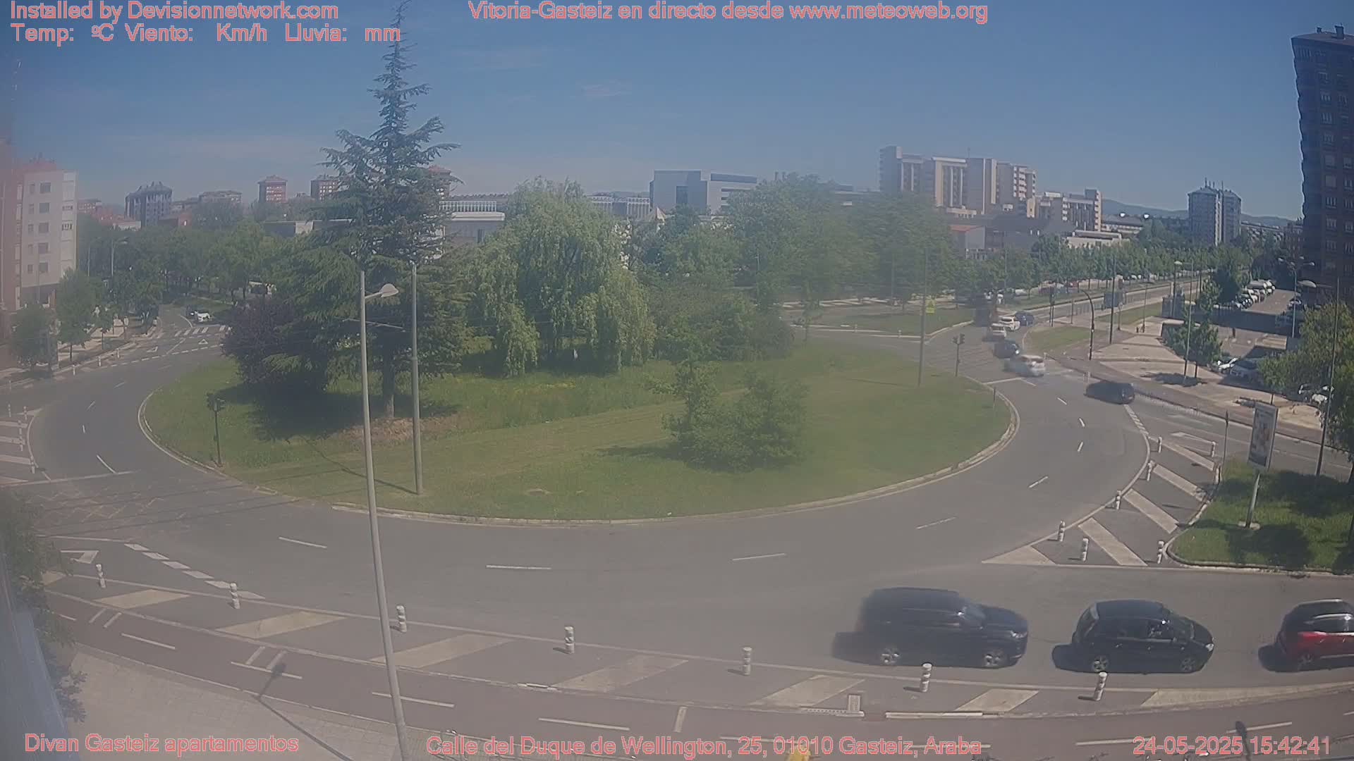 A sunny day reveals a roundabout surrounded by green spaces and buildings, with several cars navigating the roadways.