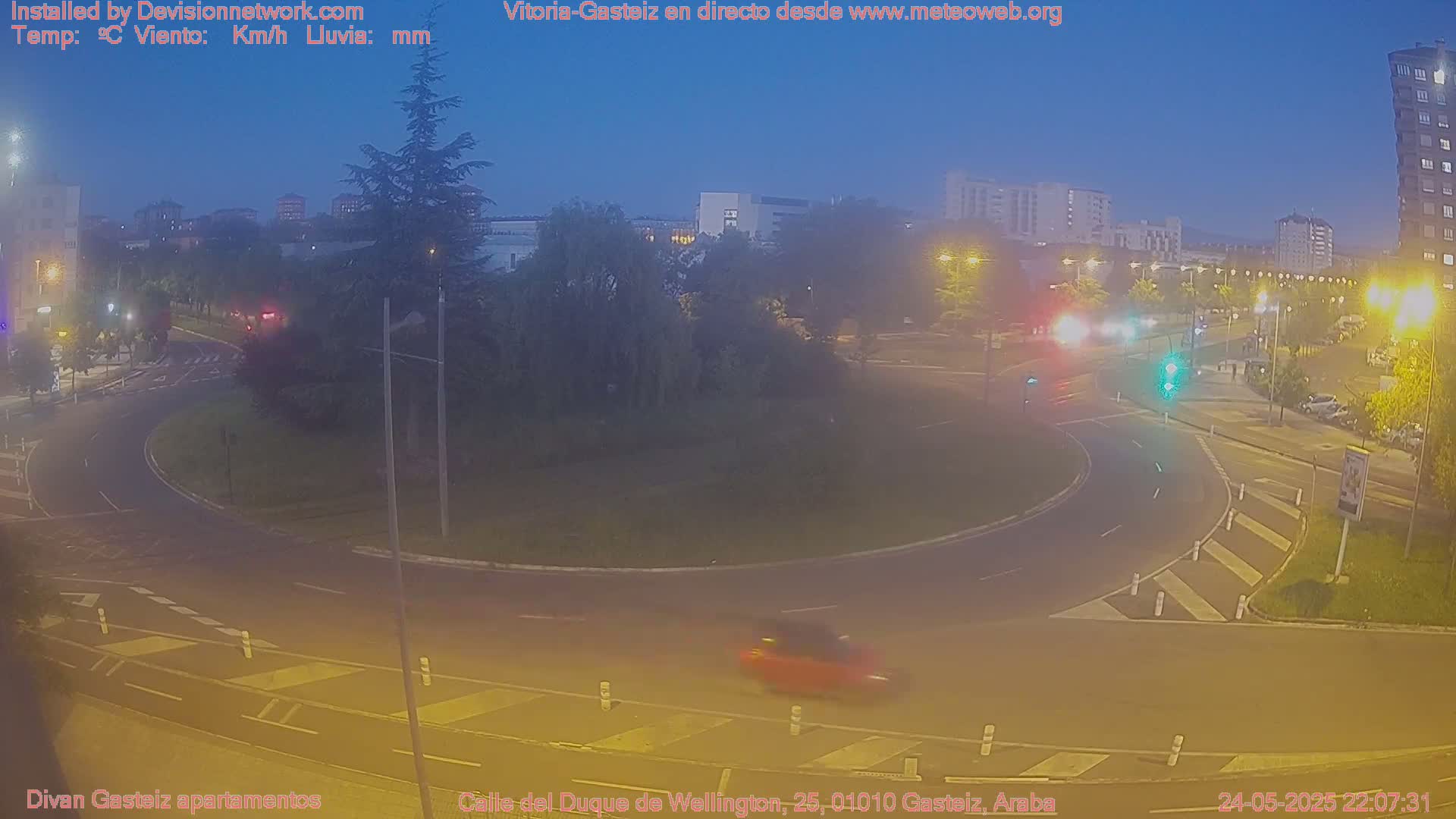 A nighttime, dimly lit view of a roundabout in a city, with a few cars visible and buildings in the background under a clear, dark blue sky.