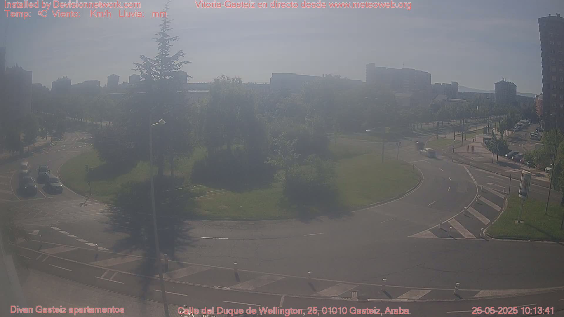 A hazy, sunny day shows a roundabout surrounded by trees and buildings in the distance.