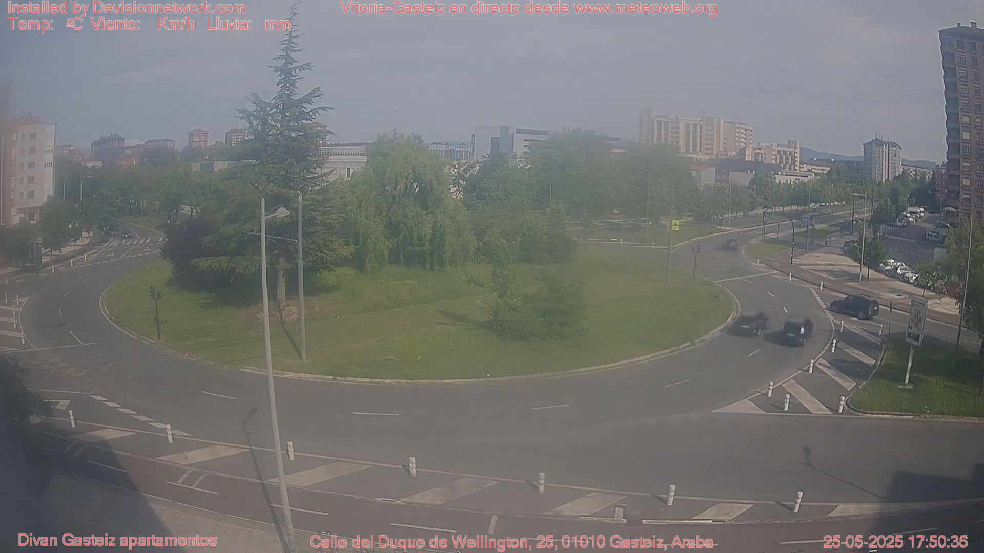 A hazy view of a roundabout and surrounding roads in a city, with several cars visible and a grassy area in the center.